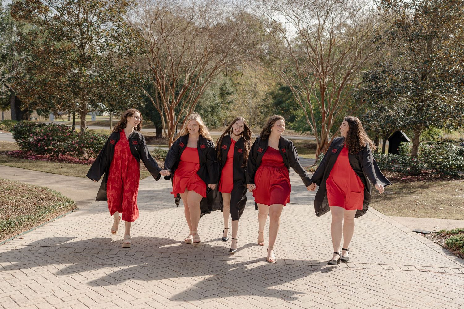 A group wearing red graduation gowns walks along a winding path through a garden setting.