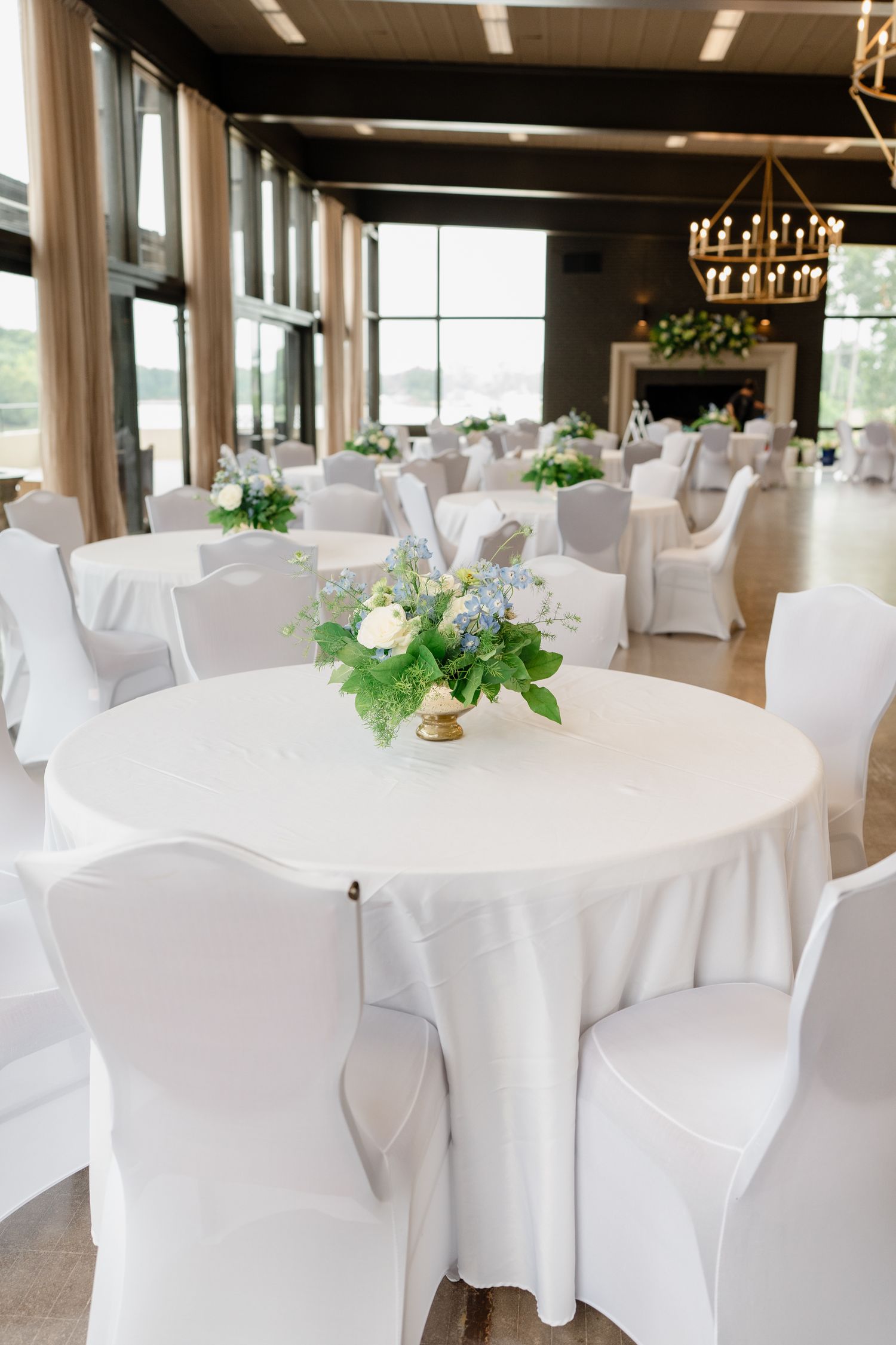 Elegant wedding reception venue with white round tables and chairs decorated with floral centerpieces in a bright room.