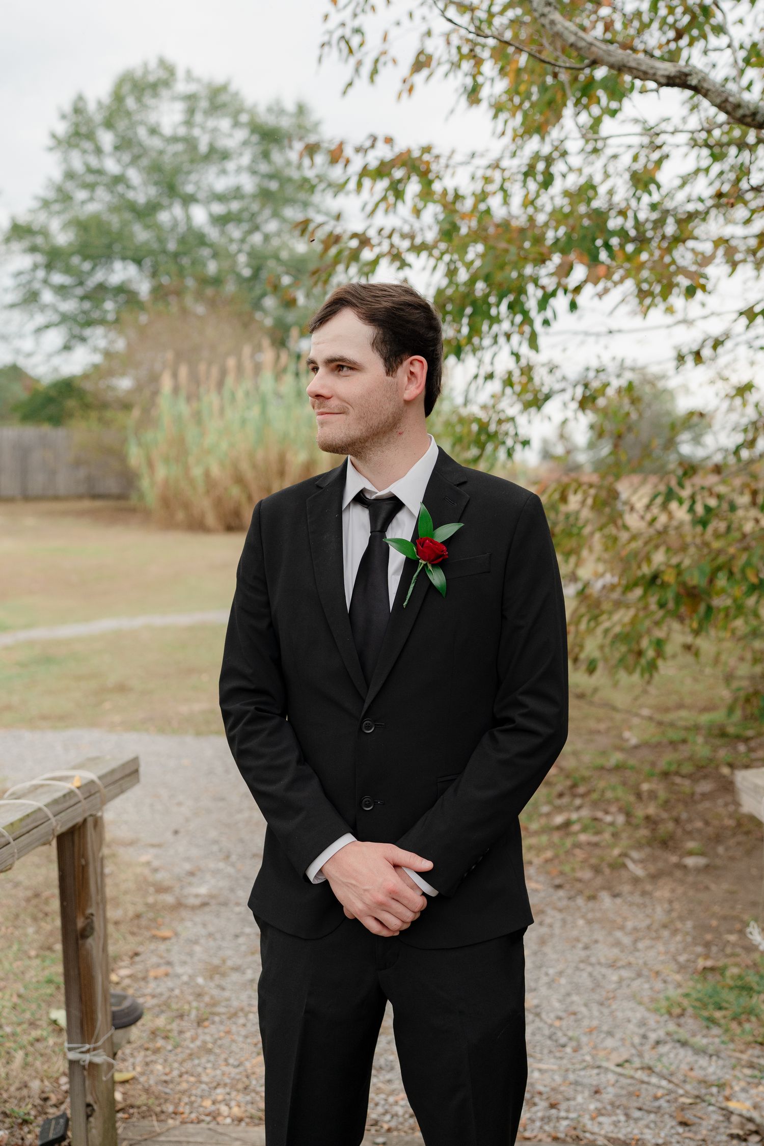 A person in a black suit with a red boutonniere stands outdoors near a wooden fence in autumn.