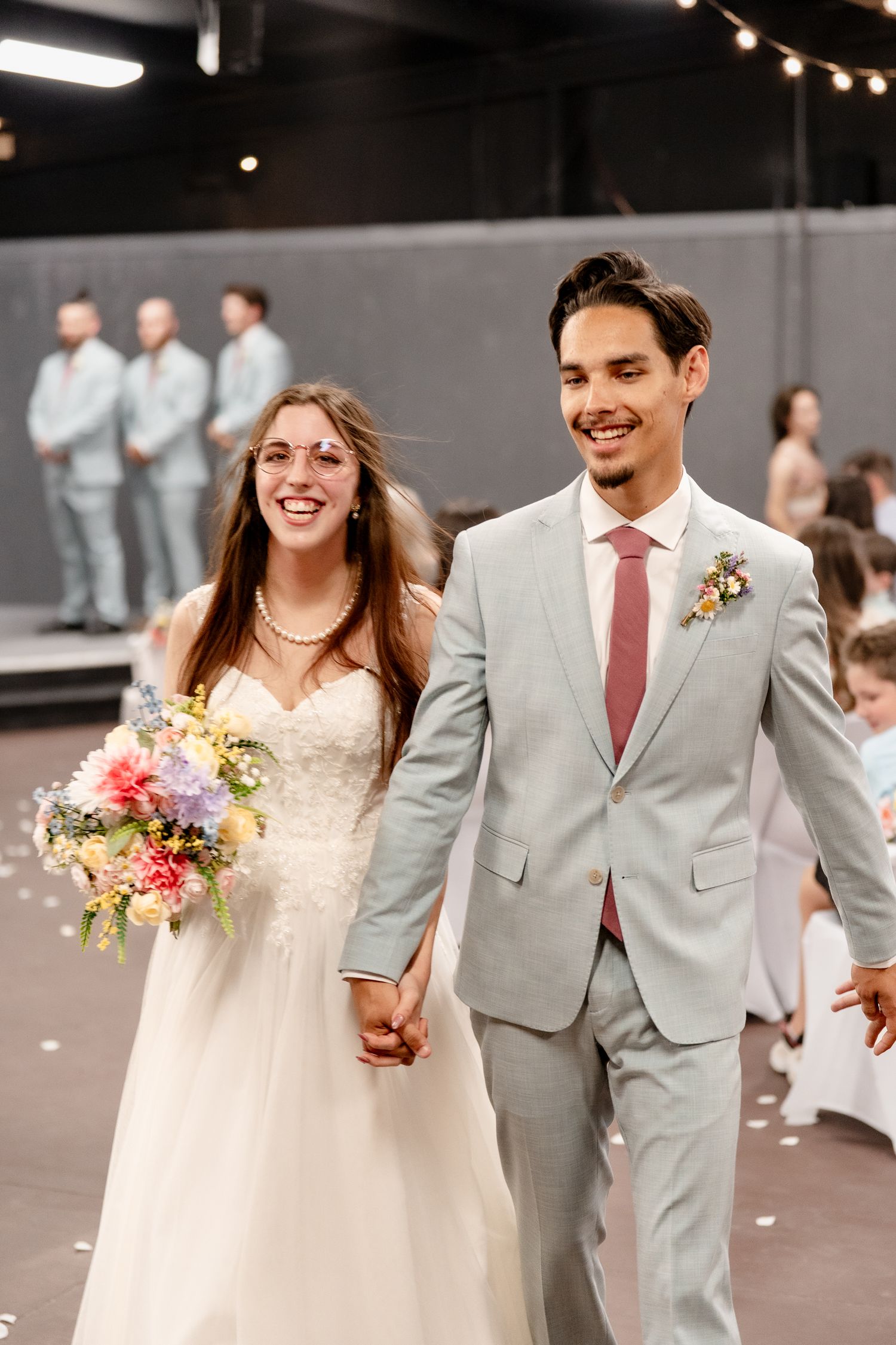A happy couple walks hand in hand at their wedding, with the bride in white and groom in a light gray suit.
