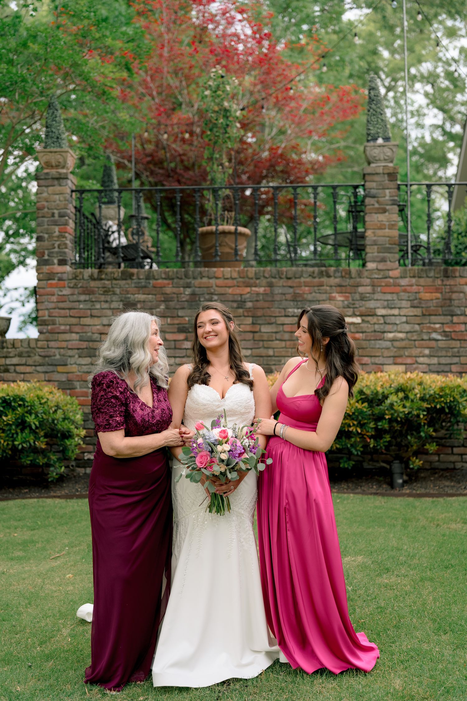 Bride in white gown poses with two bridesmaids in burgundy and pink dresses outdoors.