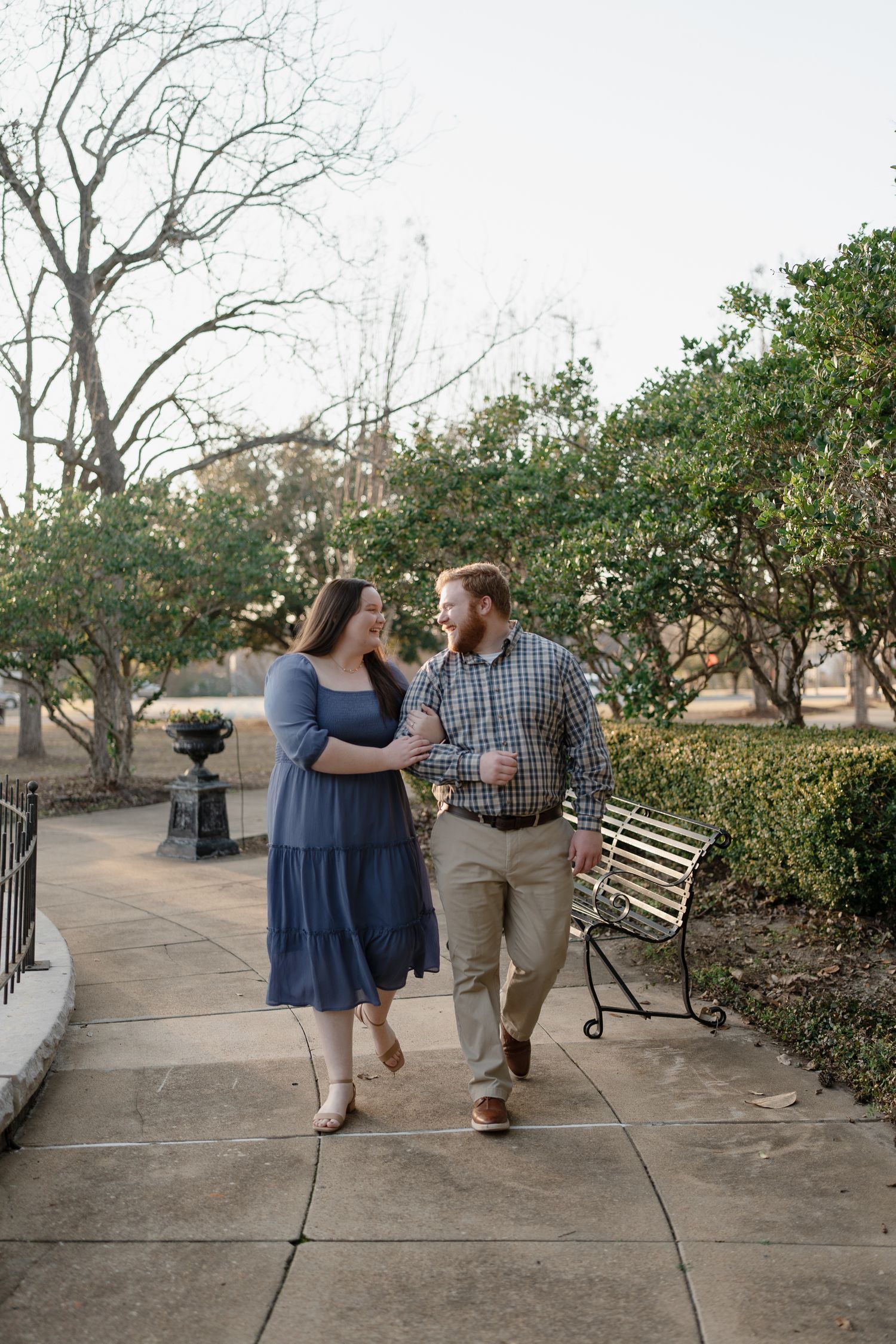 Couple walking hand in hand along a garden path, smiling at each other, surrounded by lush green hedges.