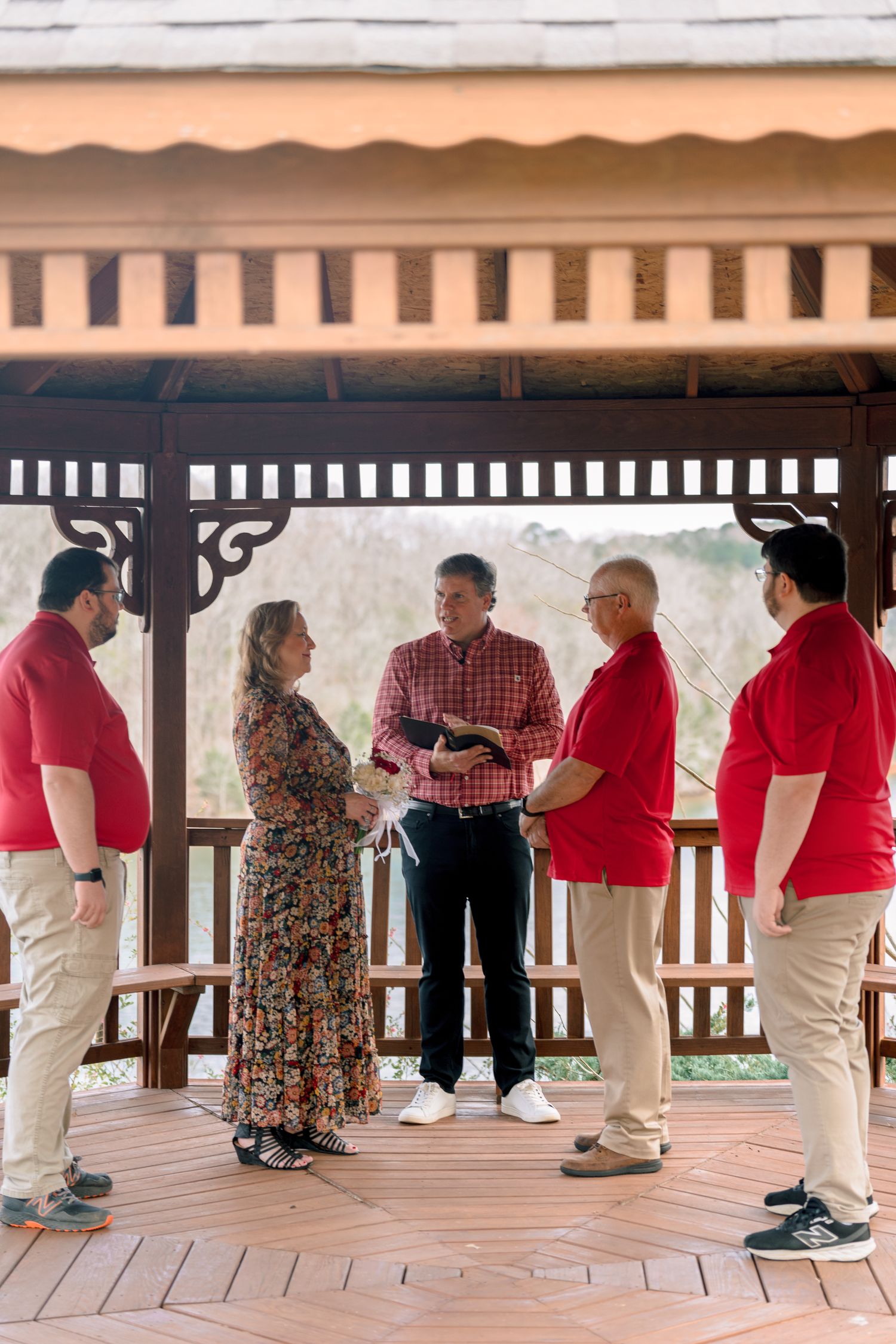 Intimate outdoor wedding ceremony in a wooden gazebo with couple and guests in red shirts.