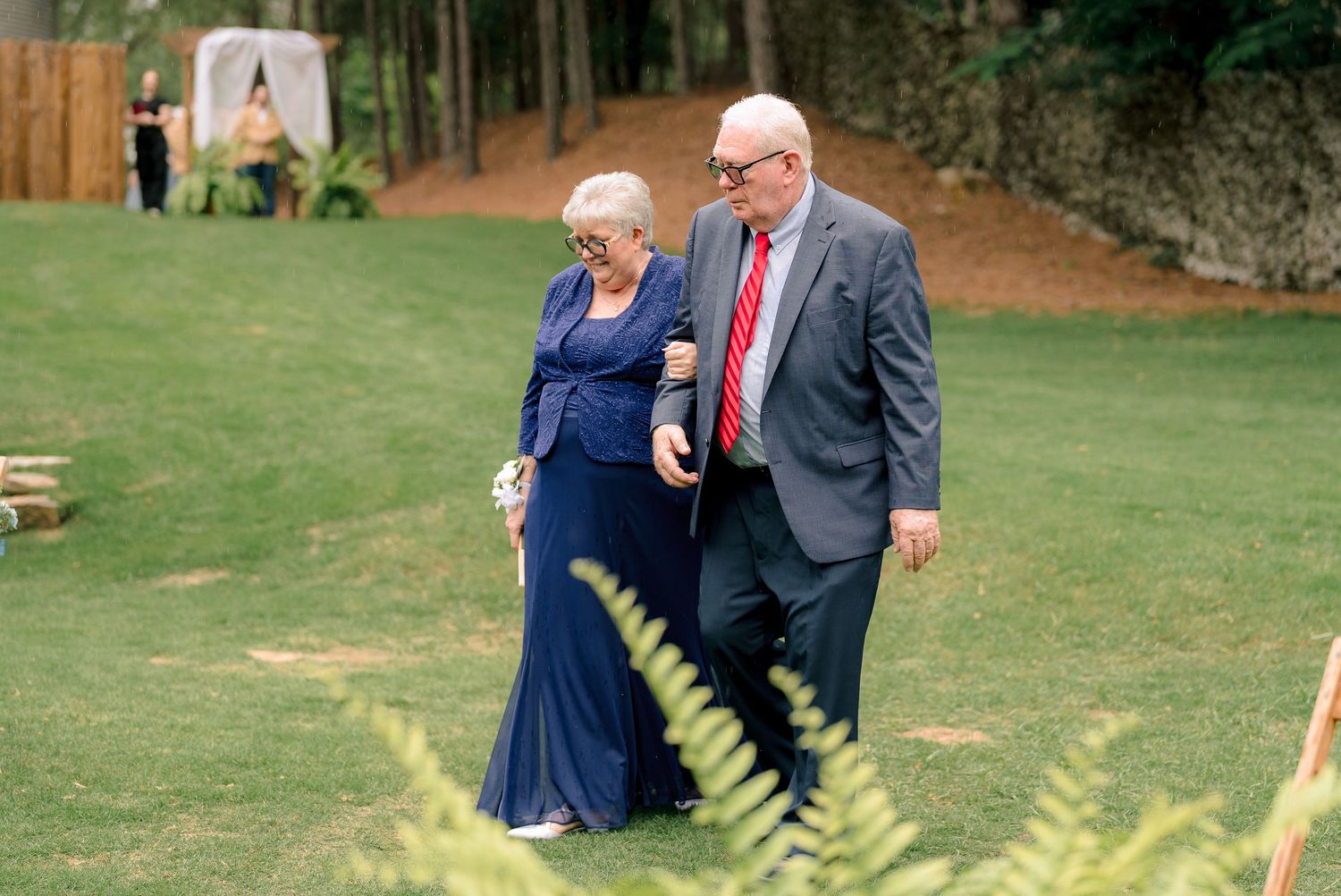 Elderly couple in formal attire walking arm-in-arm across a green lawn at a wedding.
