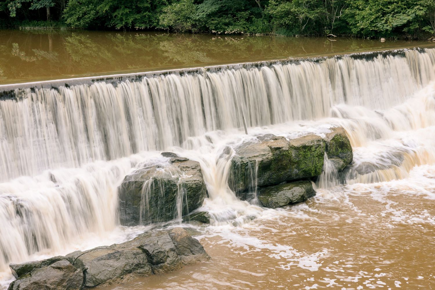A cascading waterfall flows over mossy rocks into a brown river, surrounded by lush green trees.