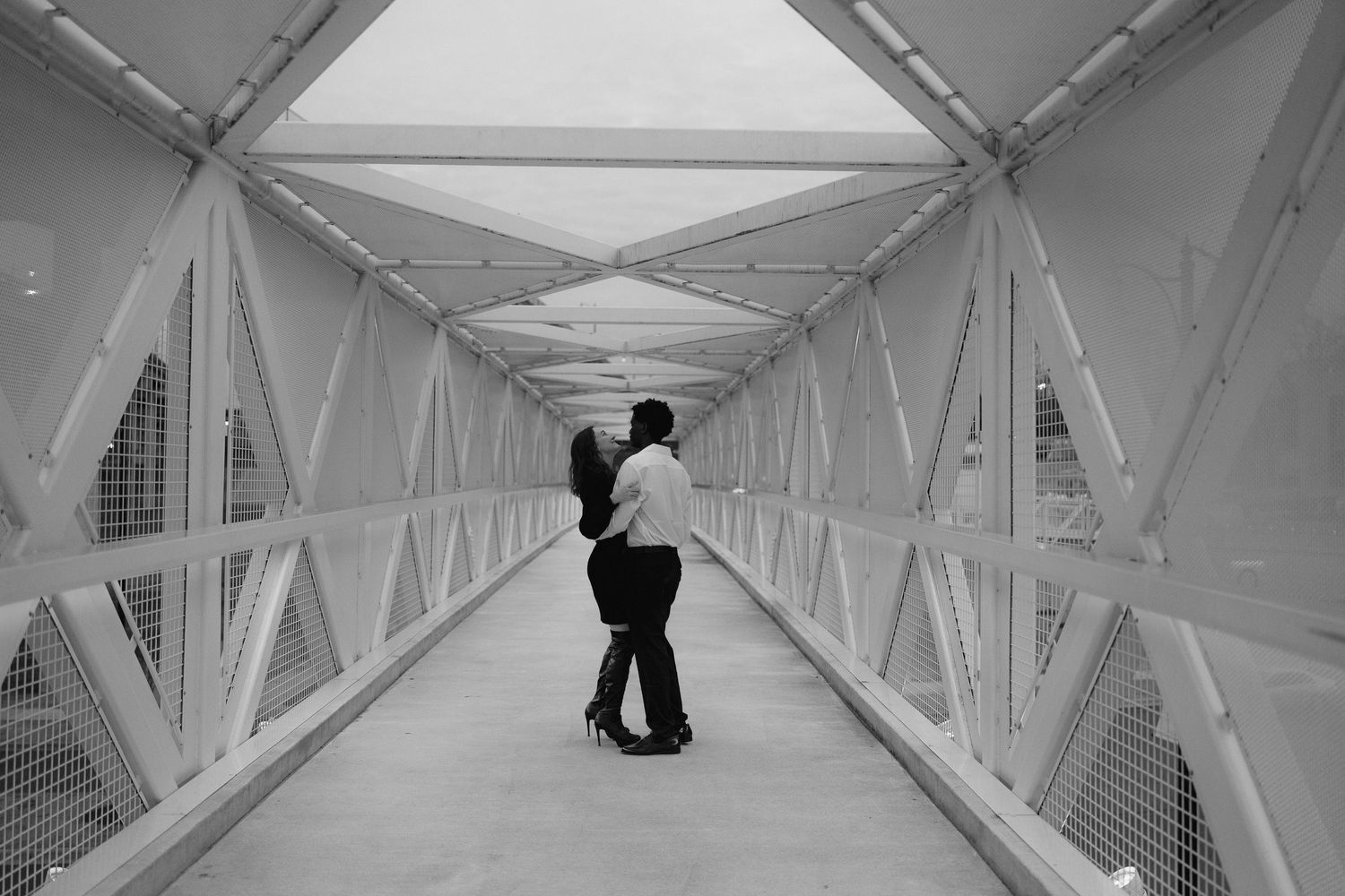 Black and white photo of a couple embracing romantically on a modern pedestrian bridge walkway.