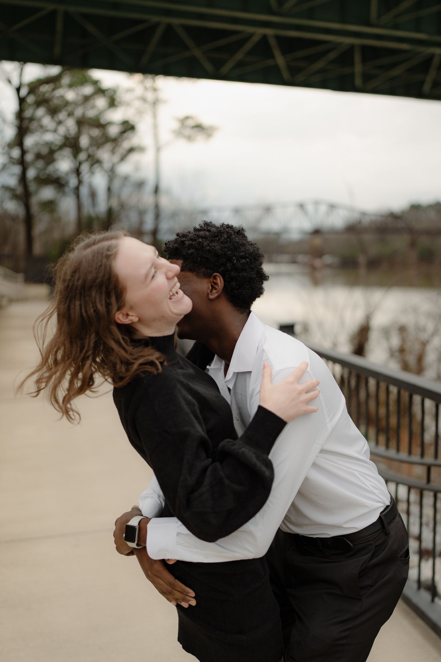 Joyful interracial couple embracing and laughing outdoors near a railing with a river in the background.
