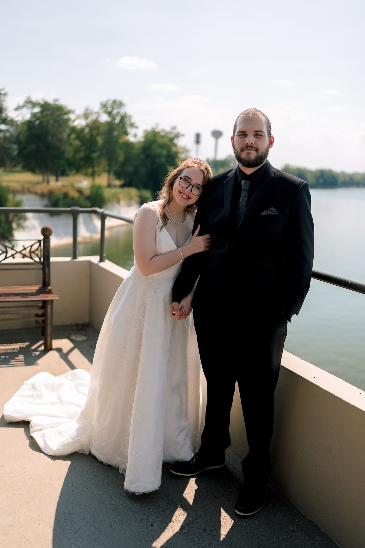 Bride and groom posing on a balcony overlooking a lake, bride in white gown, groom in black suit.