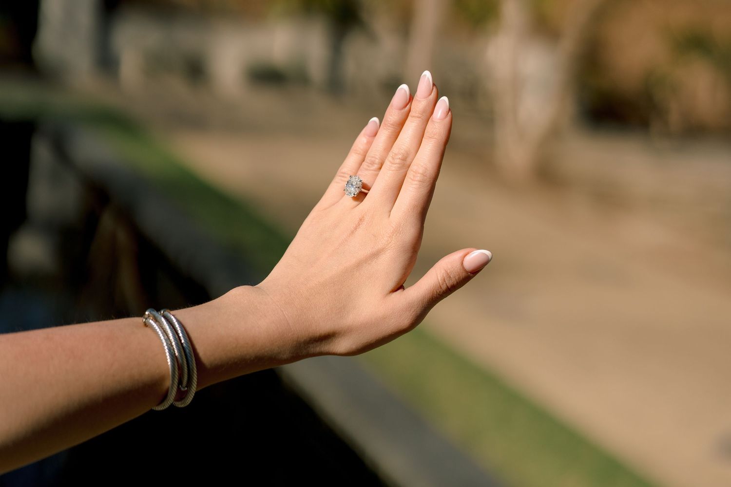 Woman displaying elegant hand with gold gemstone ring and silver bangles bracelet against blurred outdoor background.