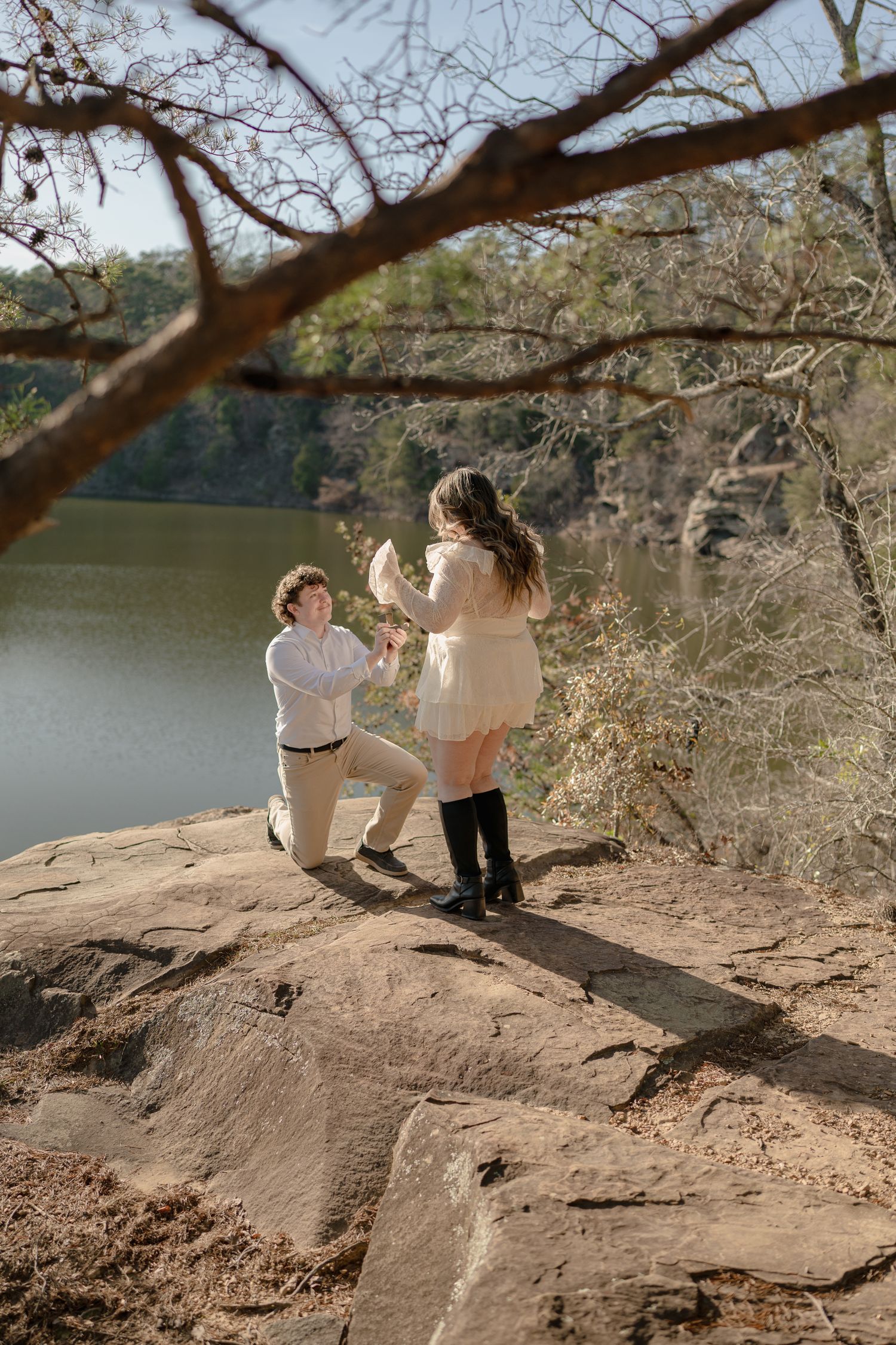 Man proposing on one knee to woman on rocky overlook above a river surrounded by trees.