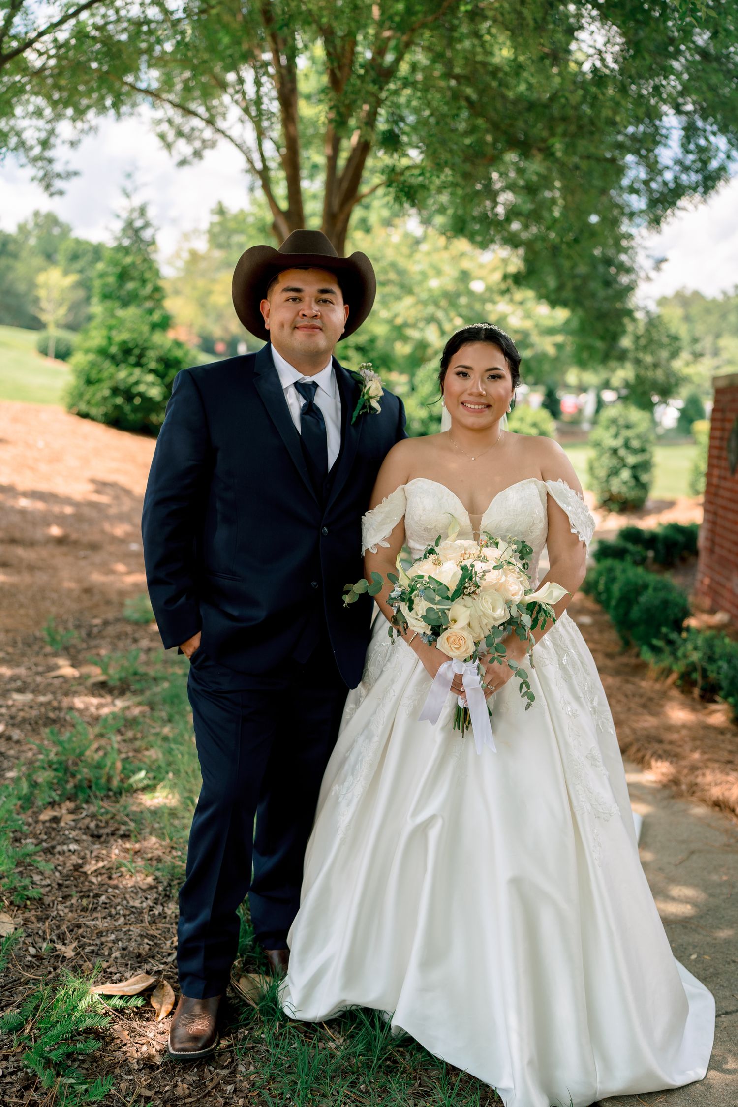 A couple poses together outdoors in formal wedding attire with lush greenery in the background.