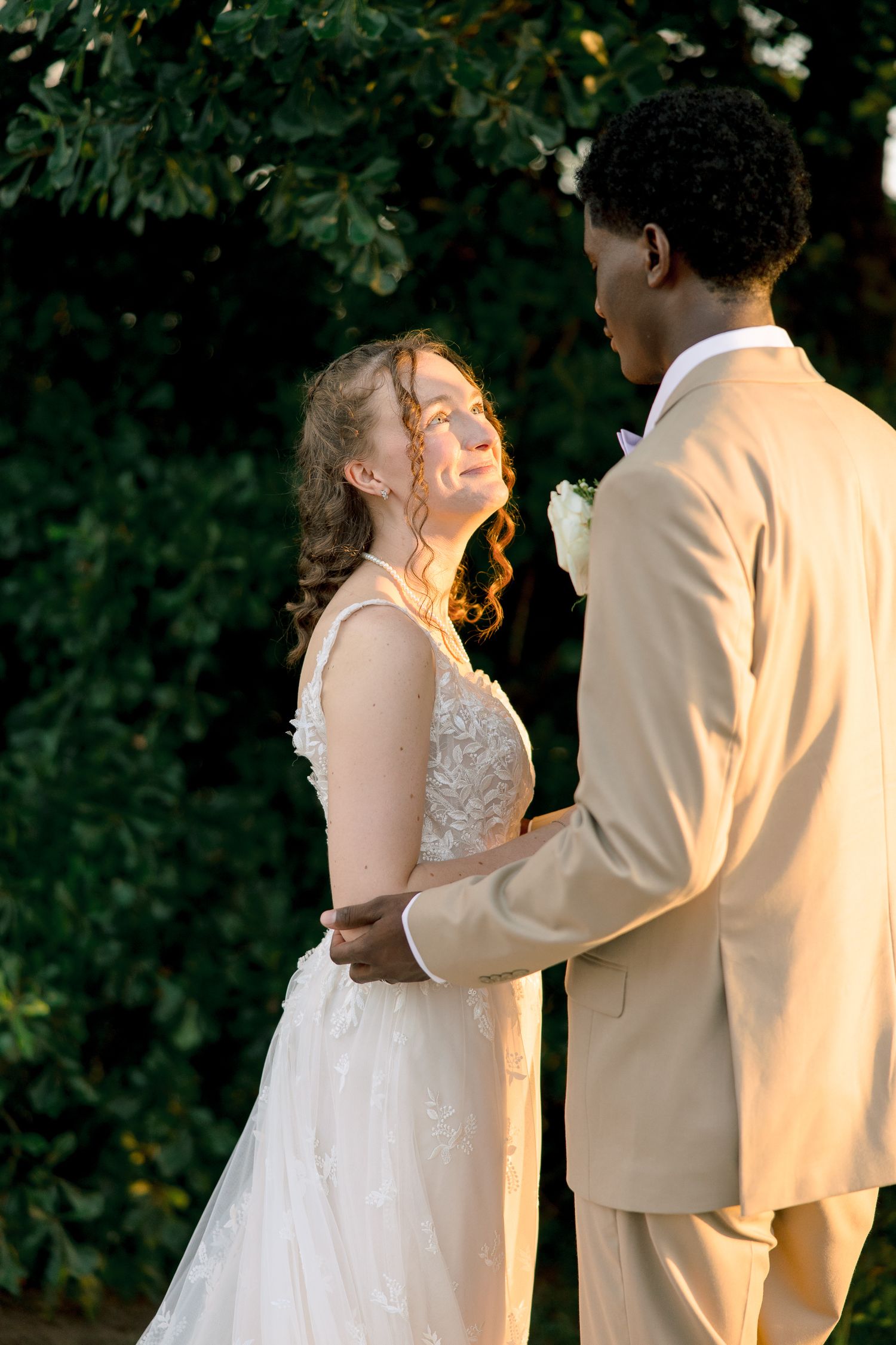 Bride in lace gown gazes up at groom in tan suit during golden hour portrait session in lush garden setting.