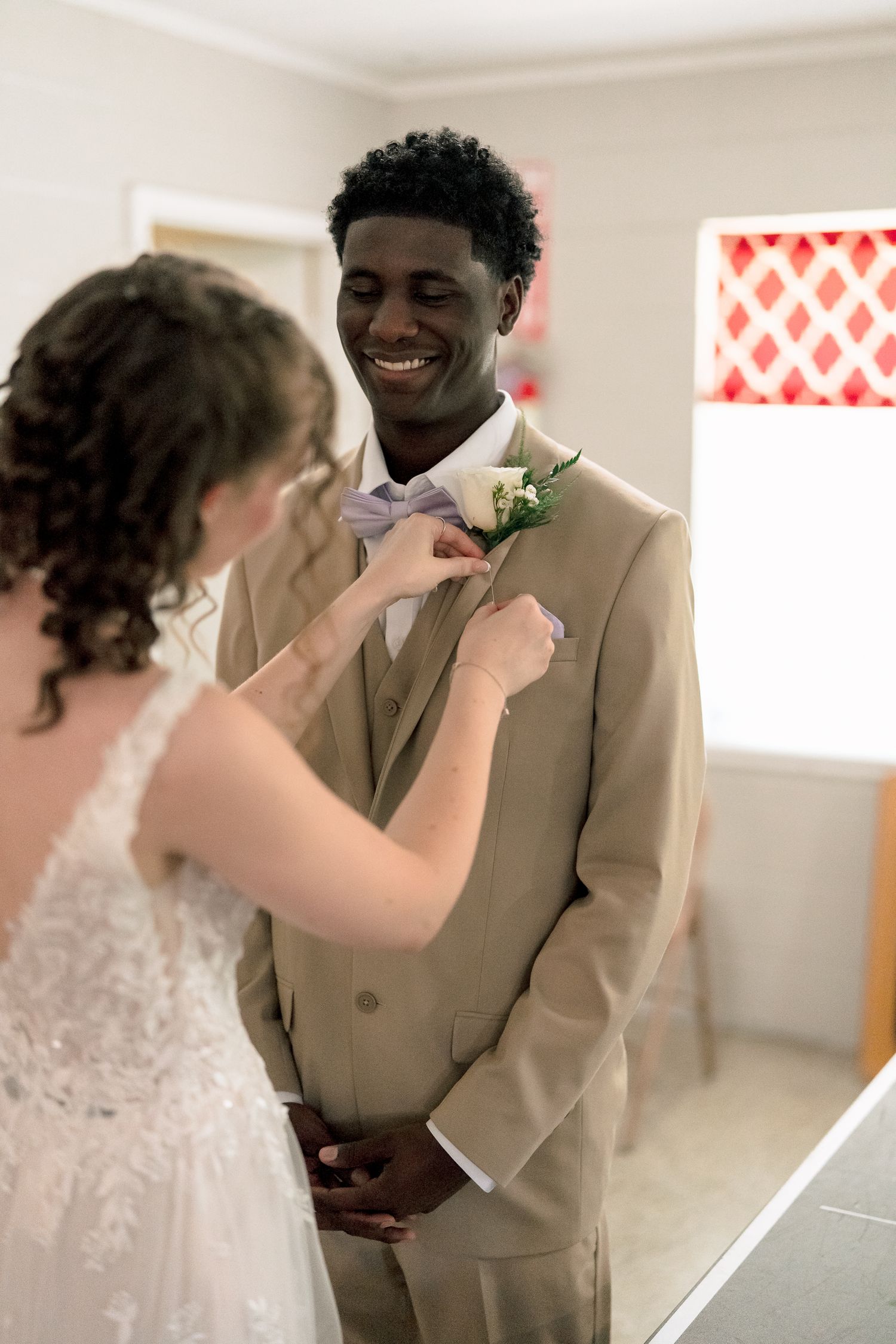 Smiling groom in tan suit getting boutonniere pinned by bride in white lace wedding dress.