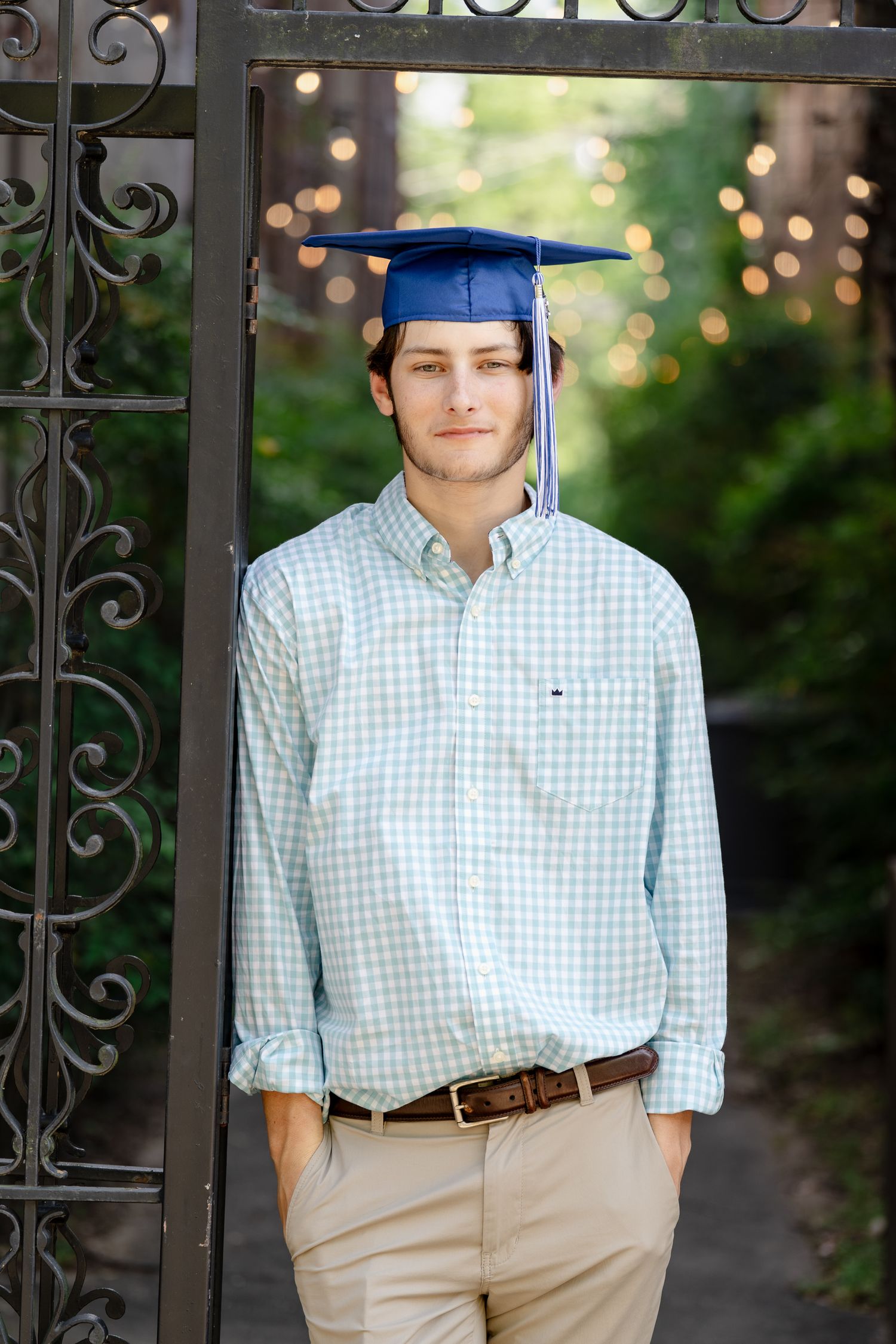 Graduate in light blue checkered shirt and blue cap poses by decorative metal gate.