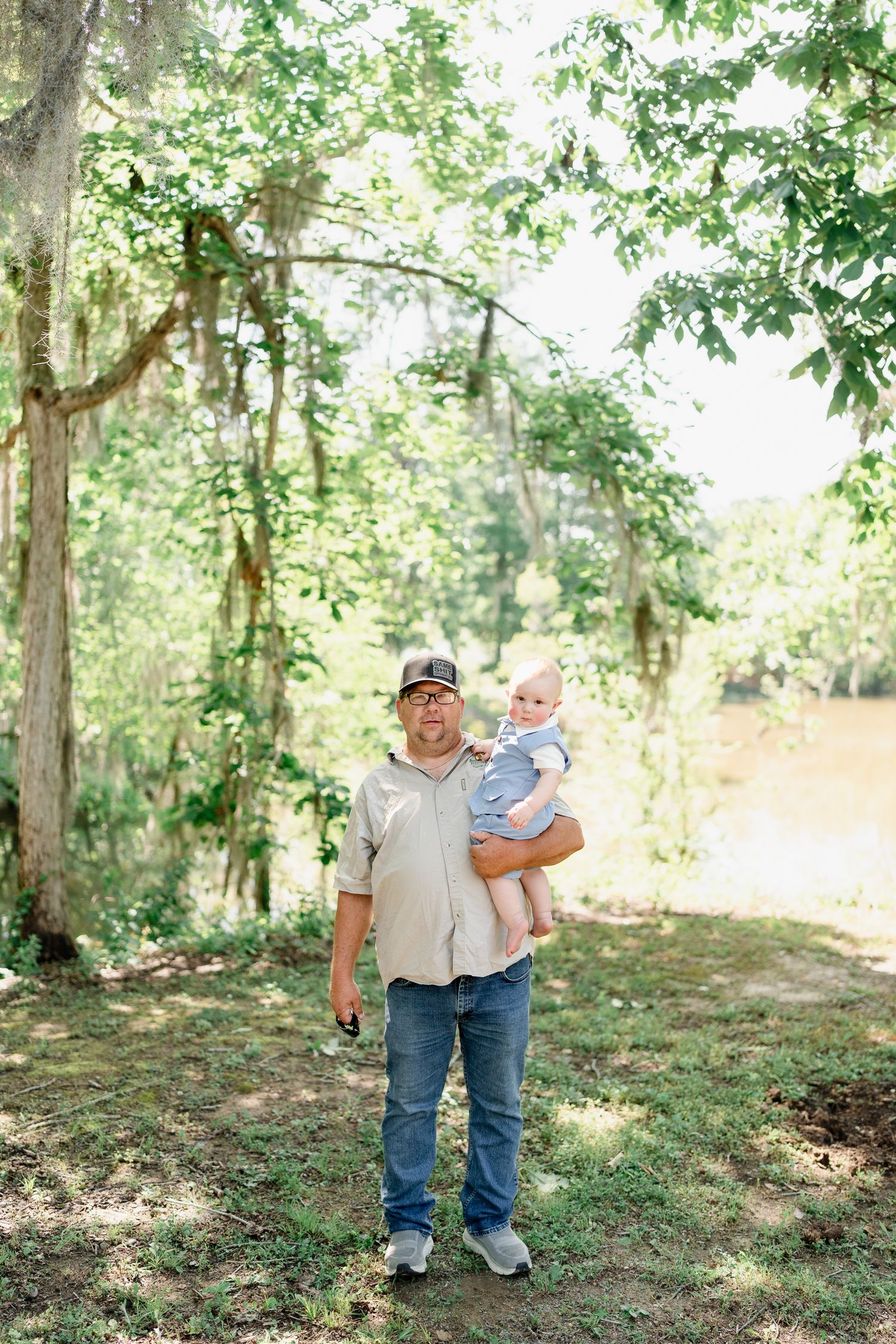 Man in casual attire holds an infant while standing in a sunlit wooded area near a river.