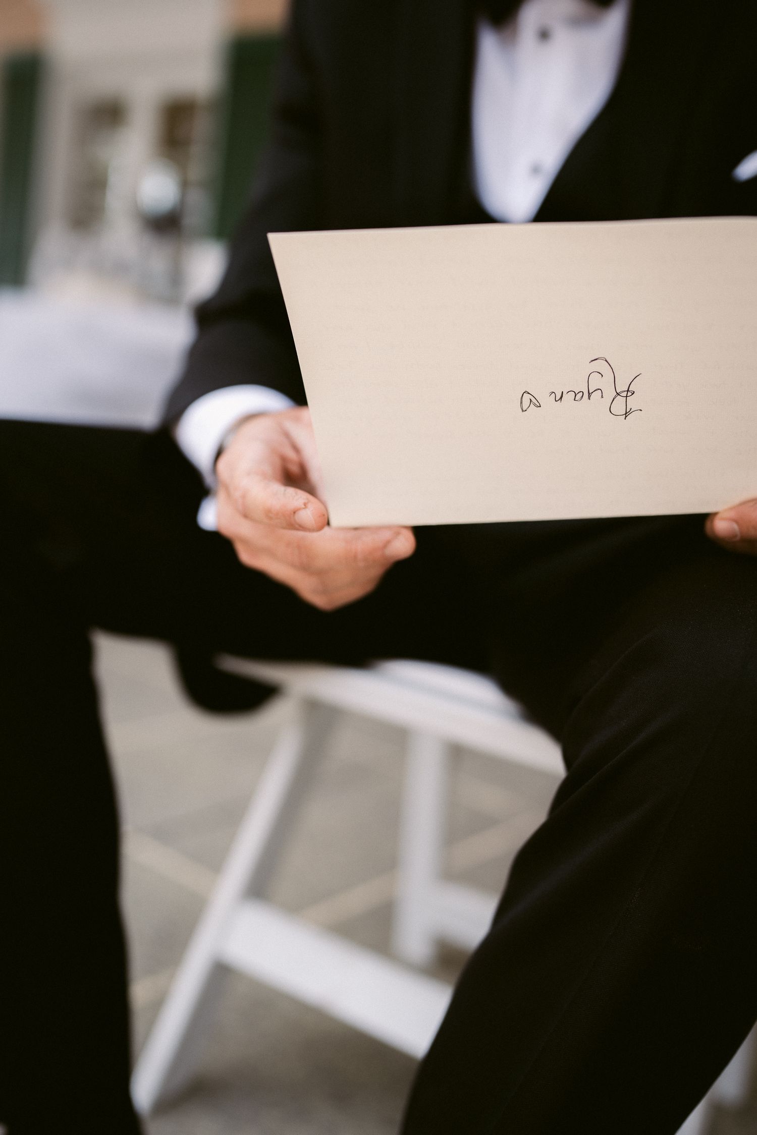 Hand in tuxedo sleeve holds a white wedding note or card.