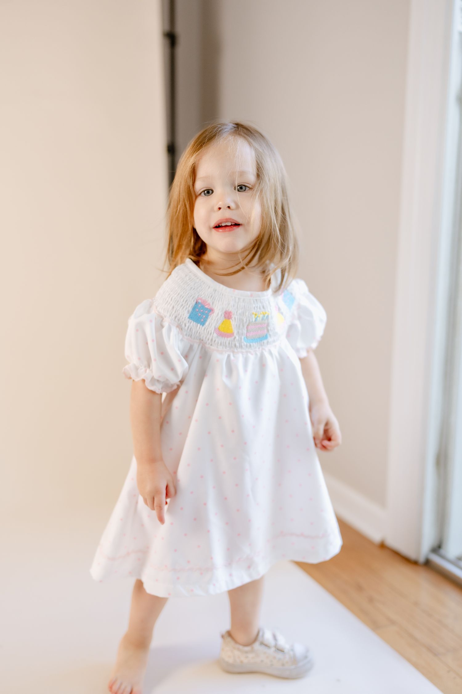 Child wearing a white dress twirls playfully in a bright indoor setting.
