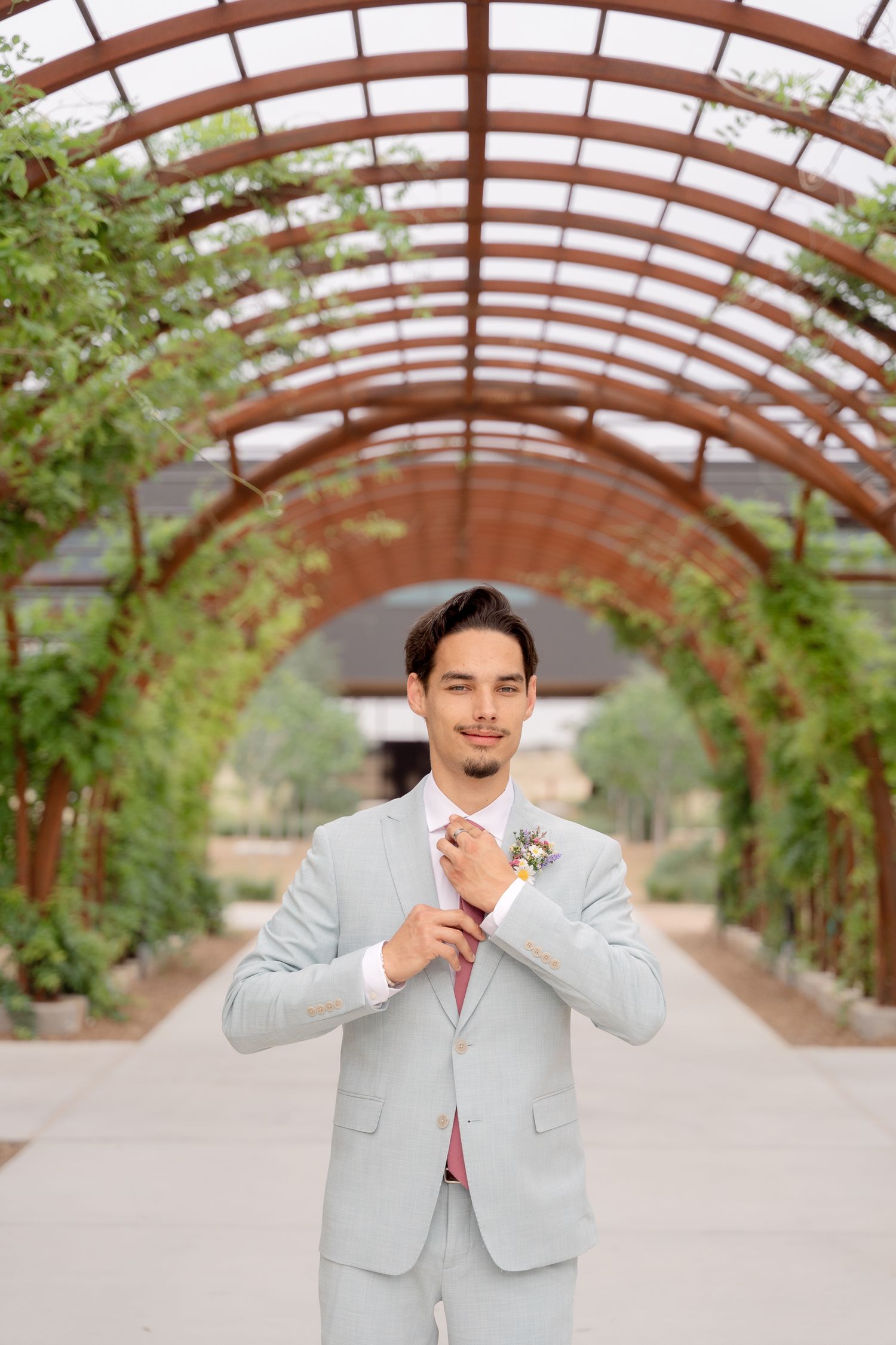 A person in a light gray suit and pink tie poses under a wooden pergola in an outdoor garden setting.