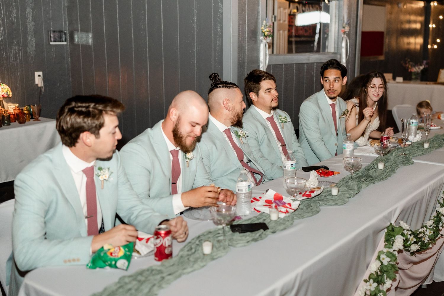 Wedding party members in mint green suits and coral ties sit at a decorated head table during reception.
