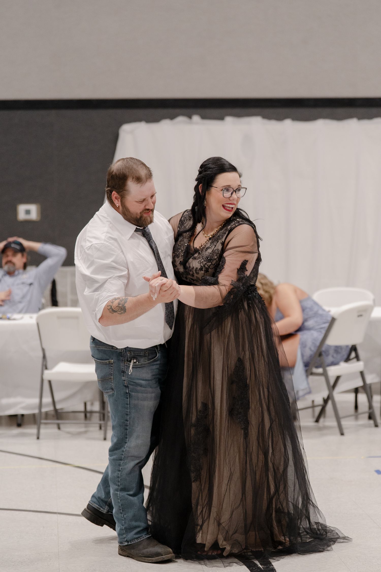A couple dancing together in a spacious room with folding chairs, the woman wearing a black tulle dress.