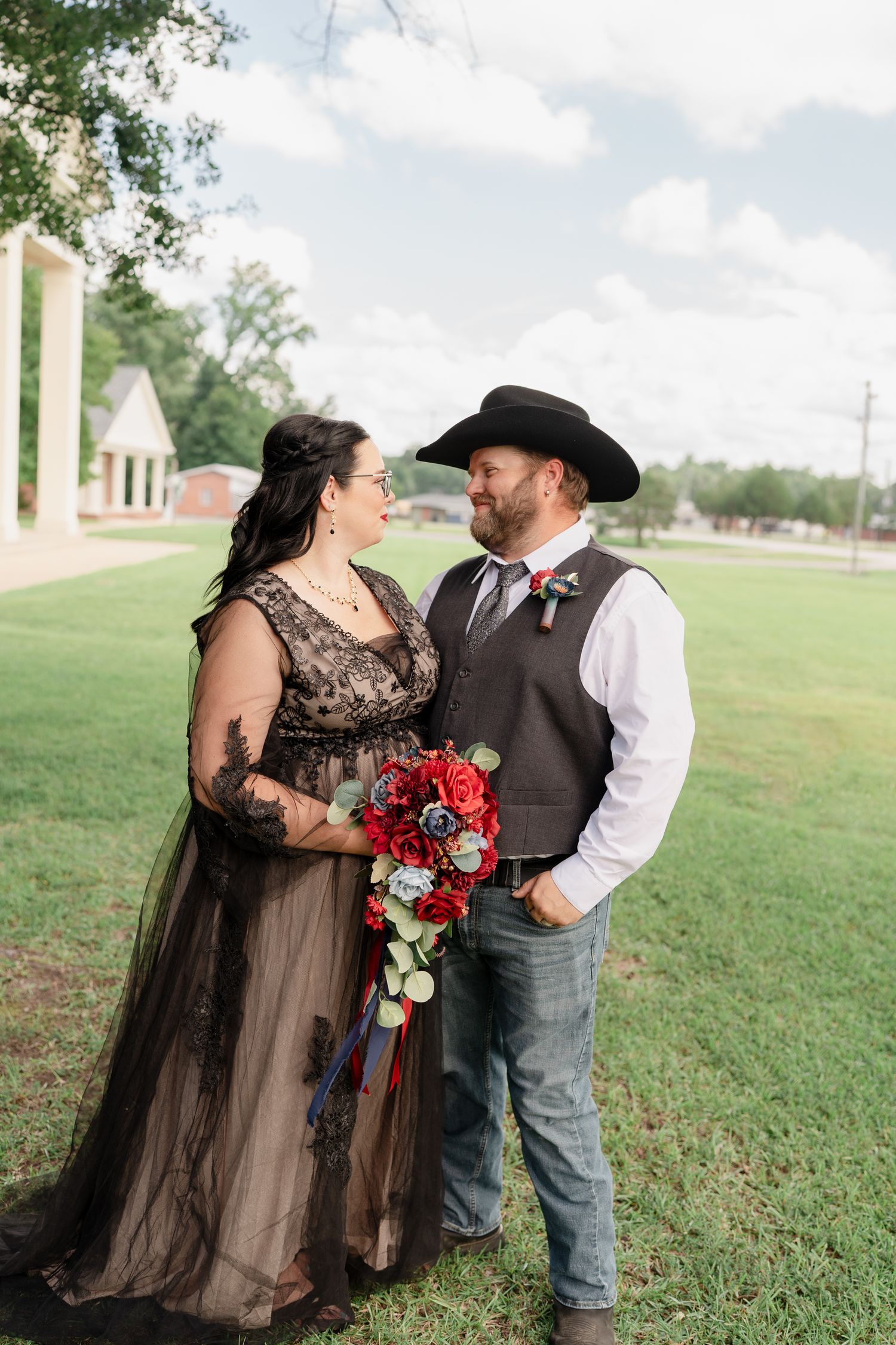 Couple in western attire poses with red bouquet on grassy lawn.