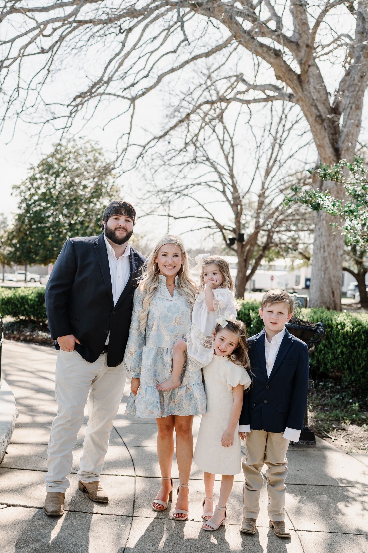 Family group poses together on sidewalk under bare winter trees in coordinated neutral outfits.