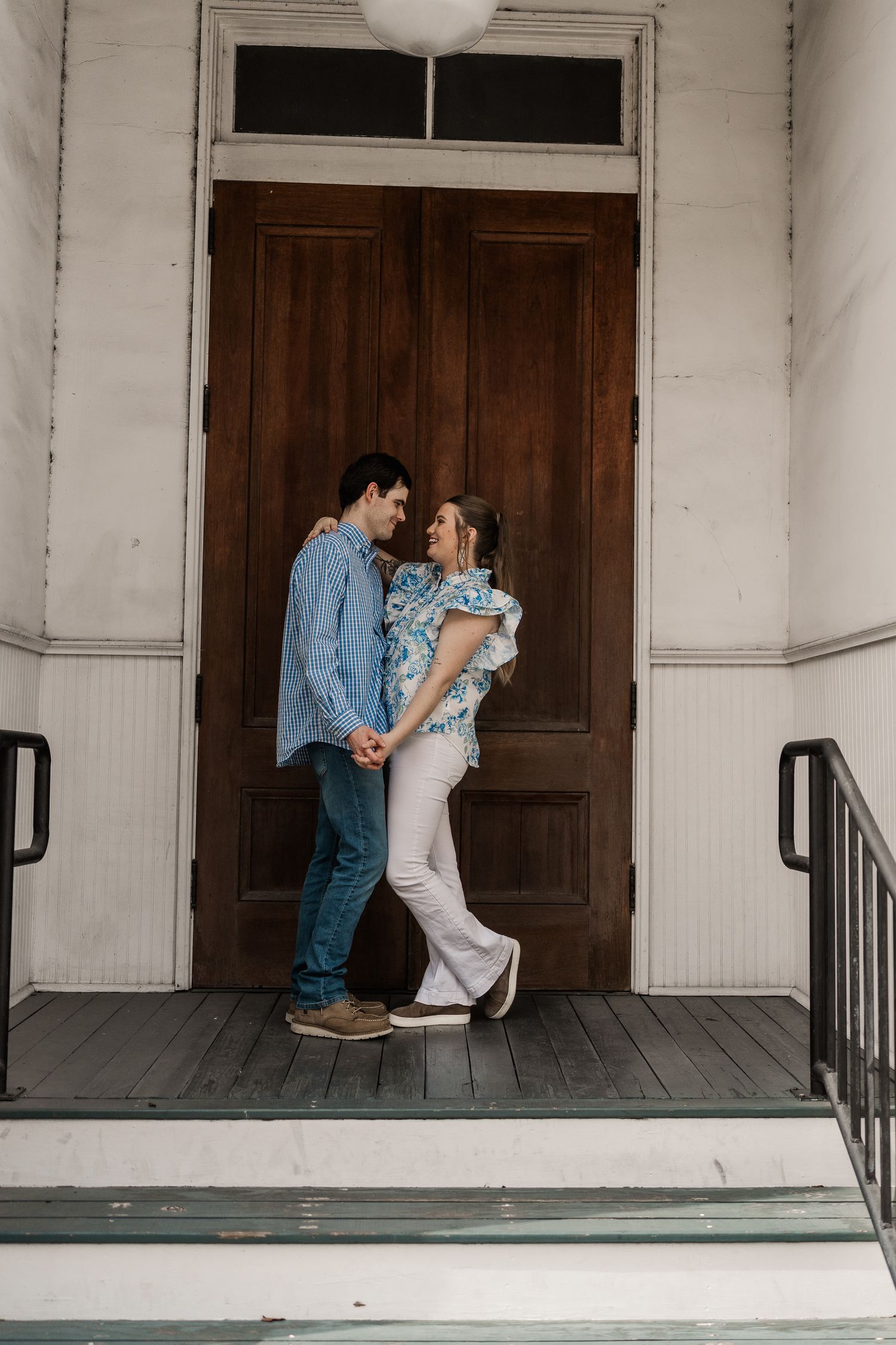 Romantic couple poses playfully on a white porch with wooden doors.