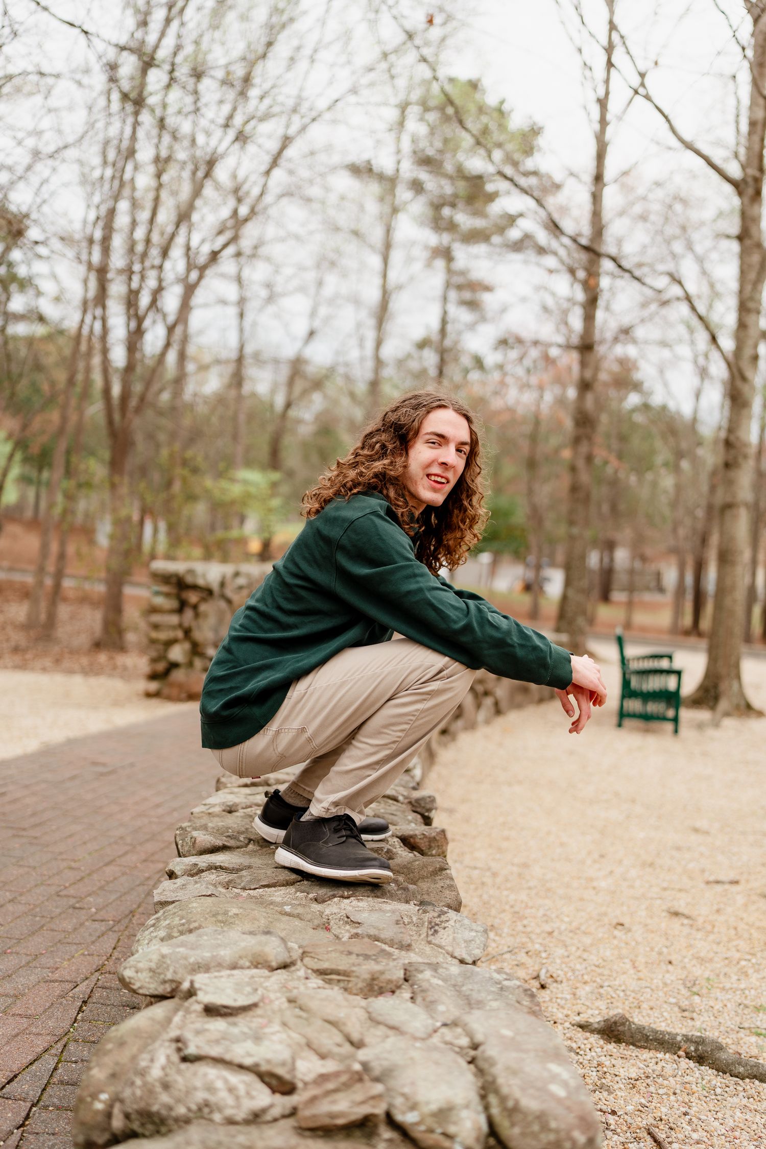 Figure crouching on stone wall wearing green sweater and khaki pants in wooded park setting.