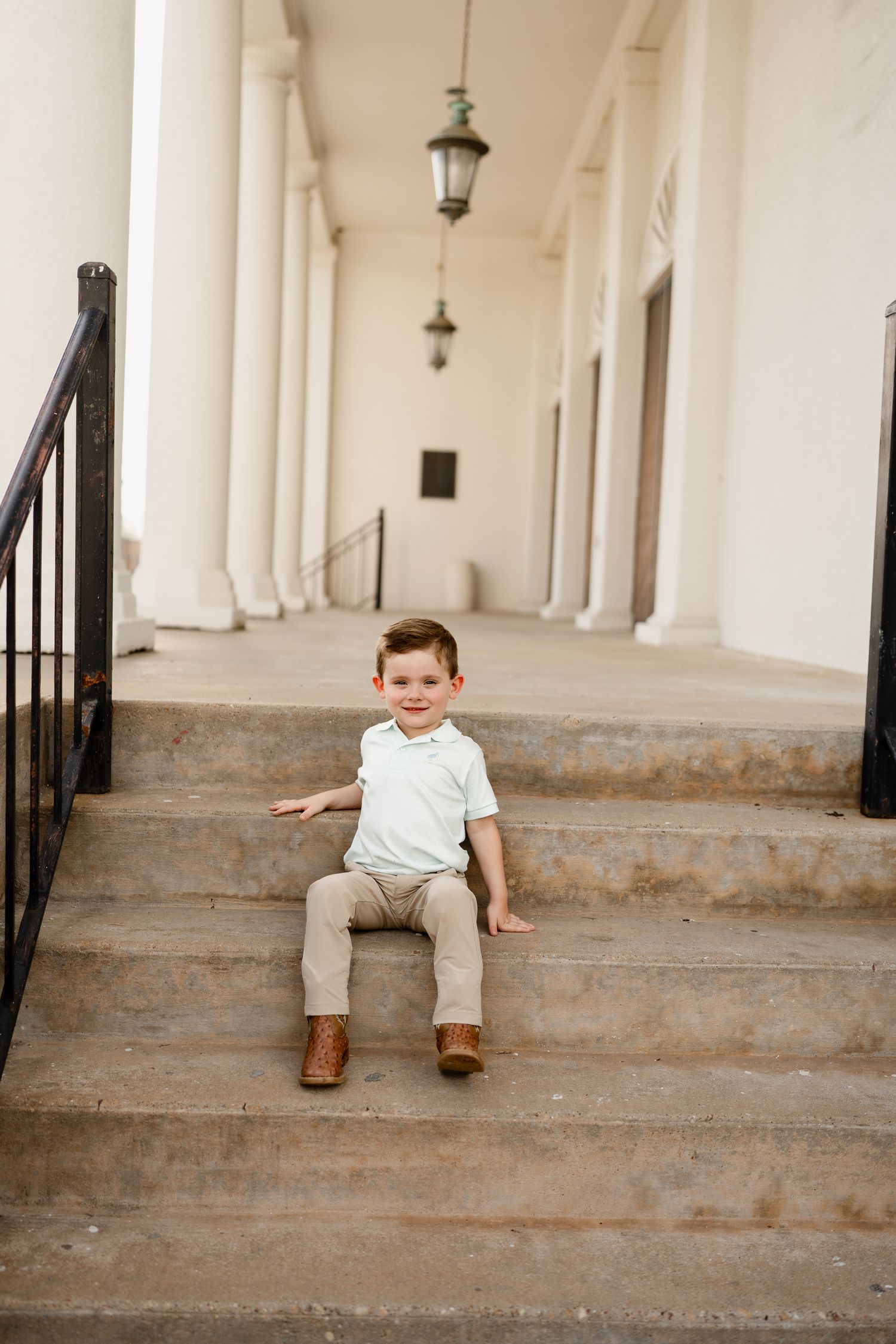 Child in white shirt and khaki pants sits on steps between white columns.