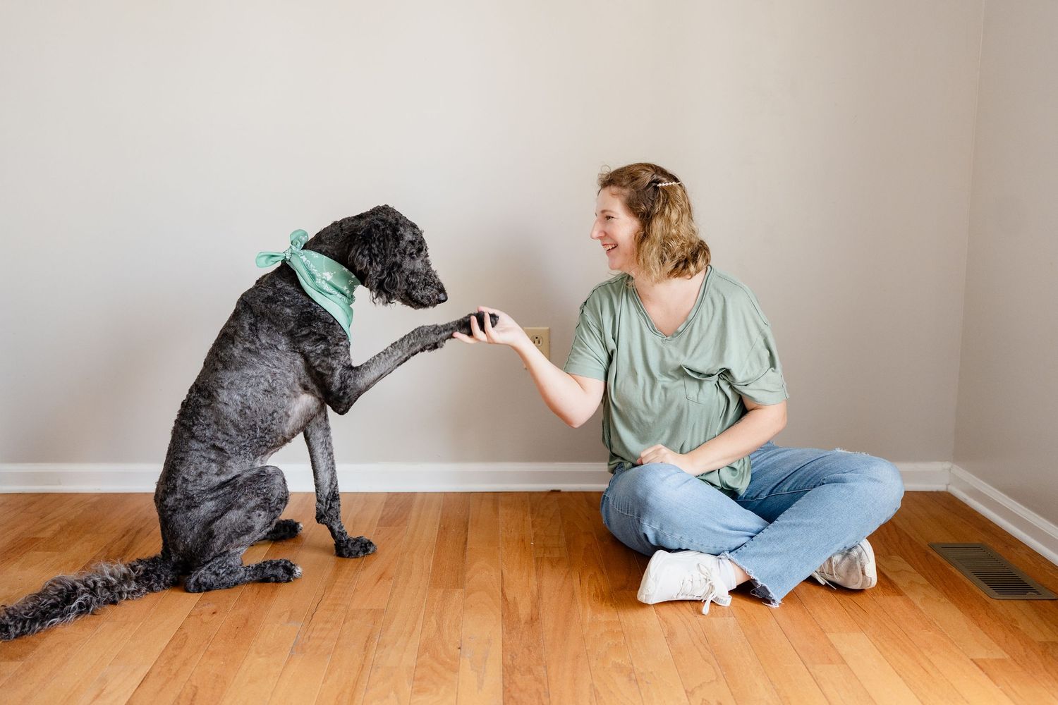 Large black dog gives high five to someone sitting cross-legged on hardwood floor while wearing mint green shirt.