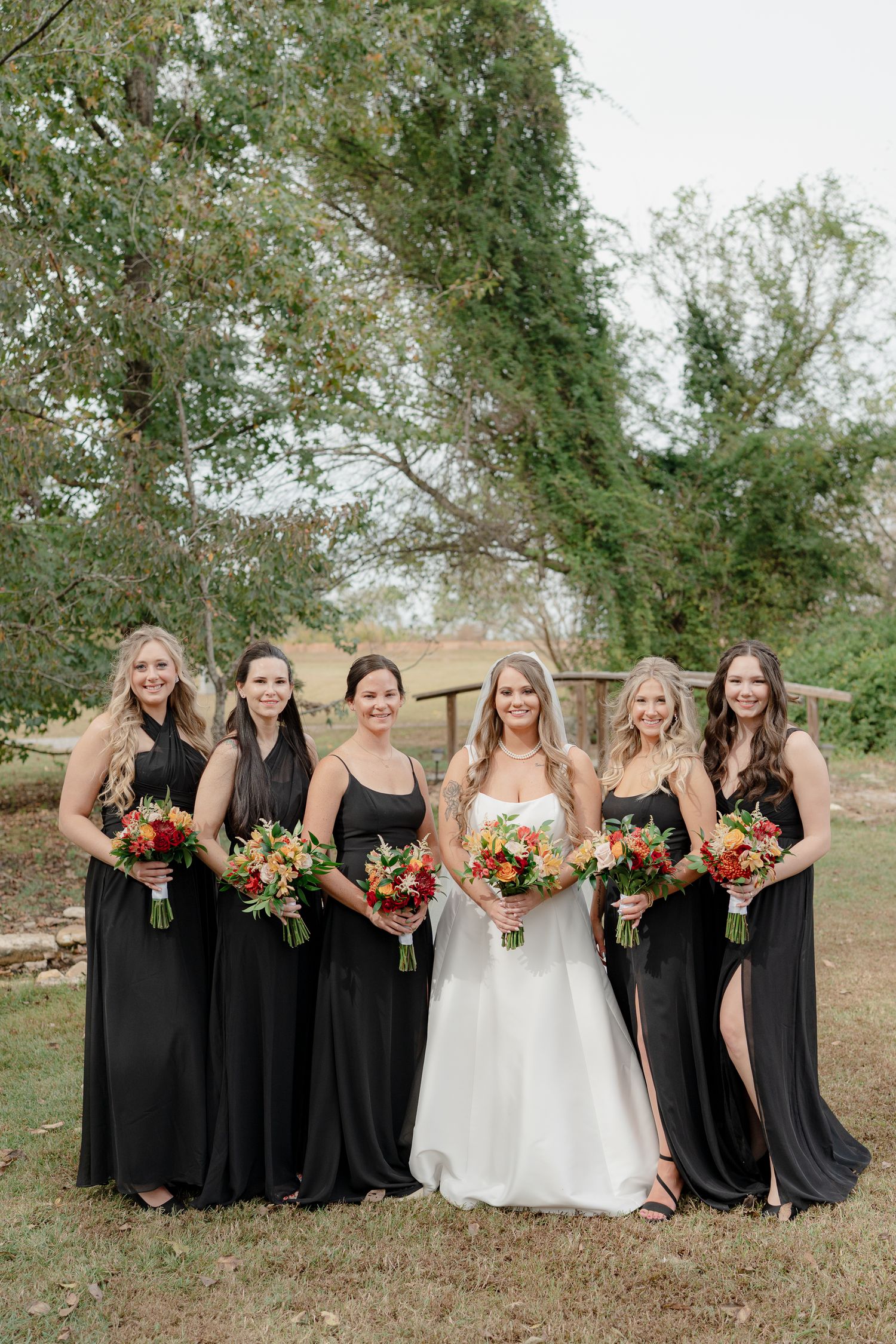 A bridal party in black bridesmaid dresses and a white wedding gown pose outdoors holding colorful fall bouquets.