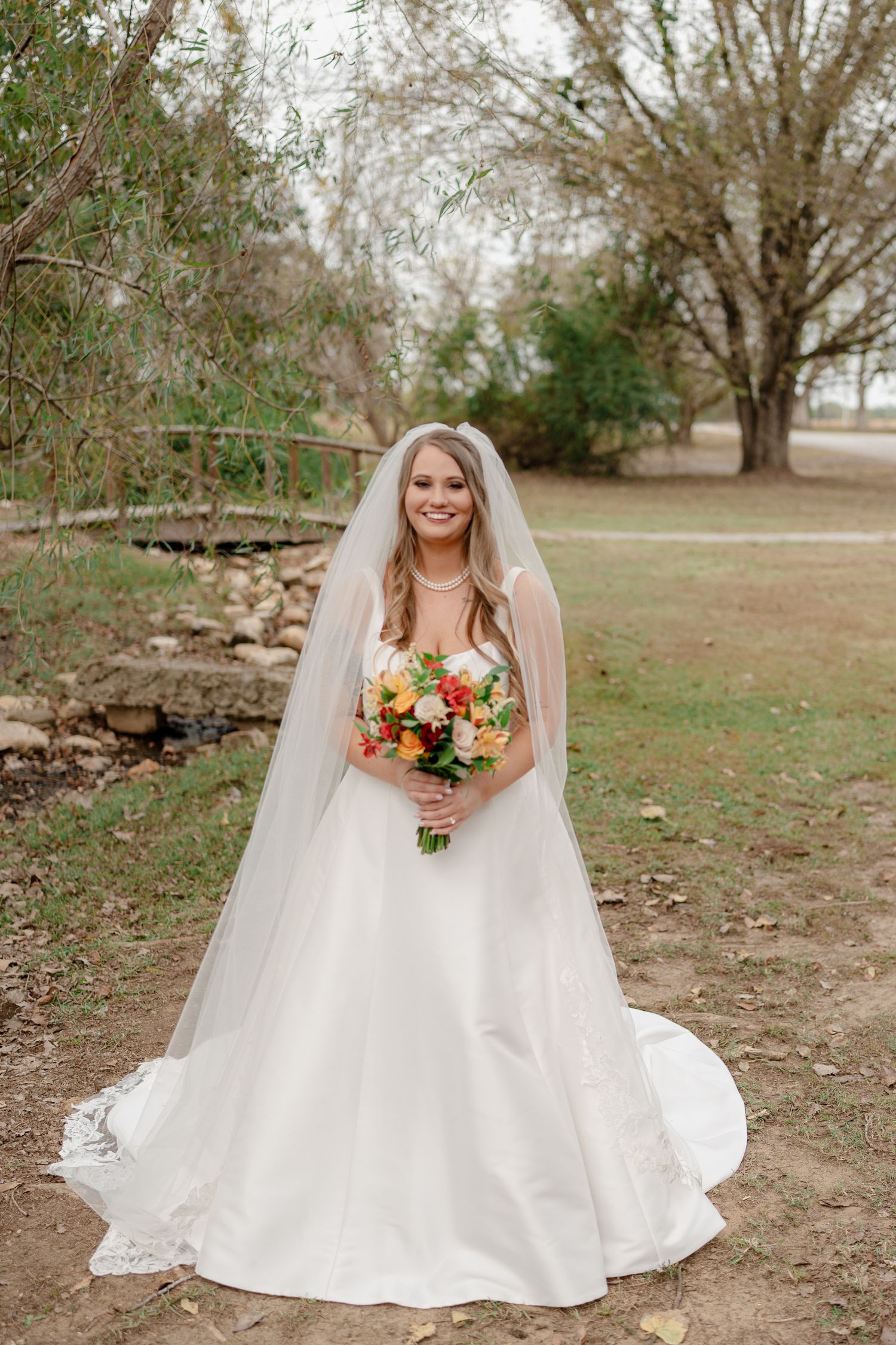 A bride in a flowing white gown sits gracefully on autumn ground holding a vibrant bouquet.