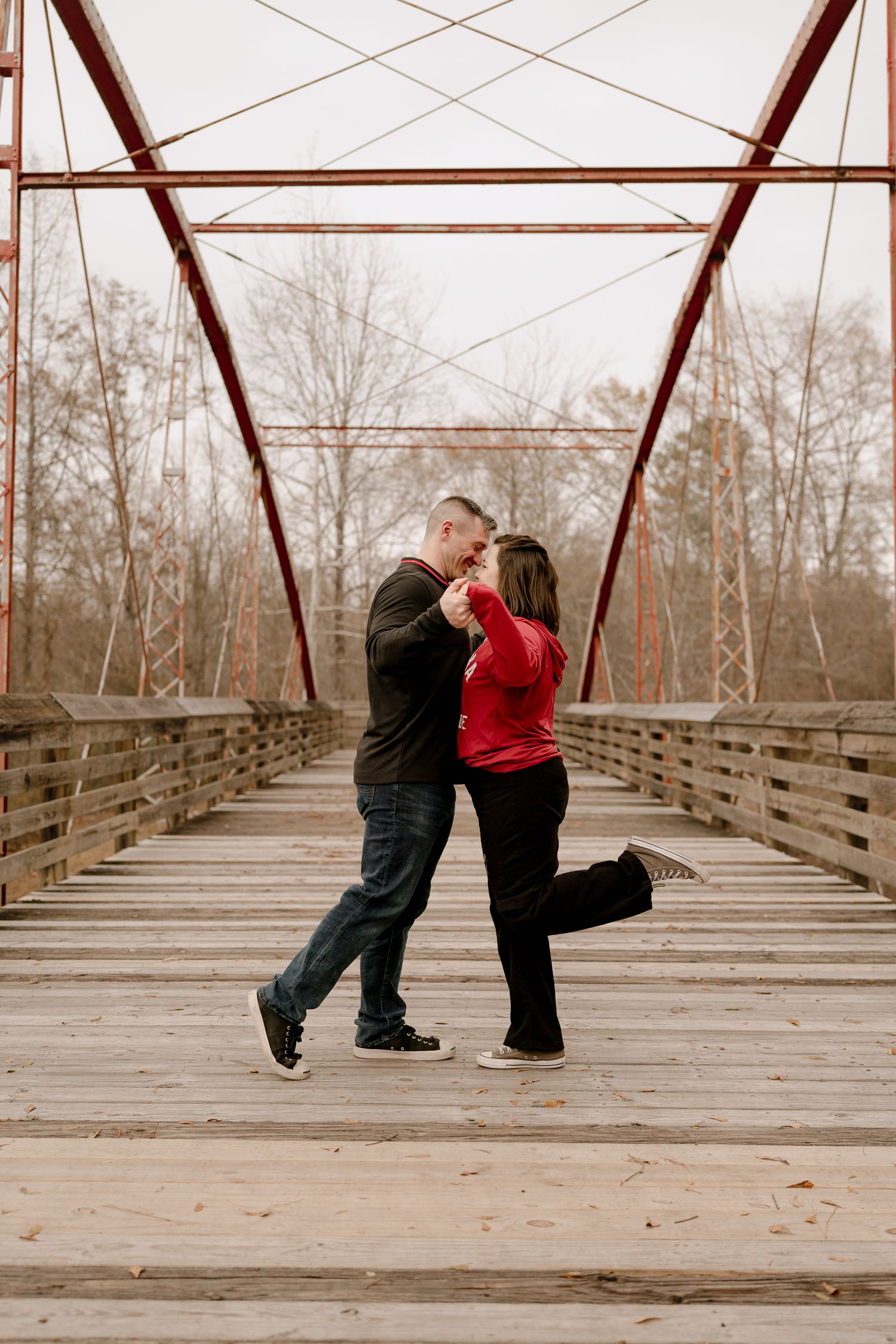 A romantic couple dancing and embracing on a rustic wooden bridge with red metal trusses.