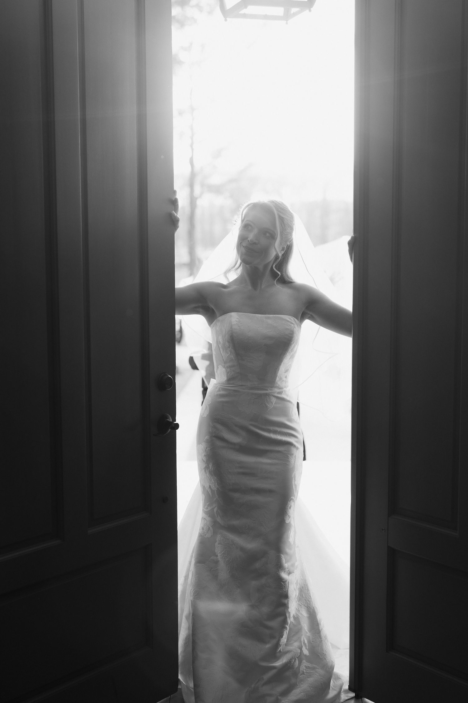 Black and white photo of a bride in a strapless gown standing in an open doorway with bright backlight.