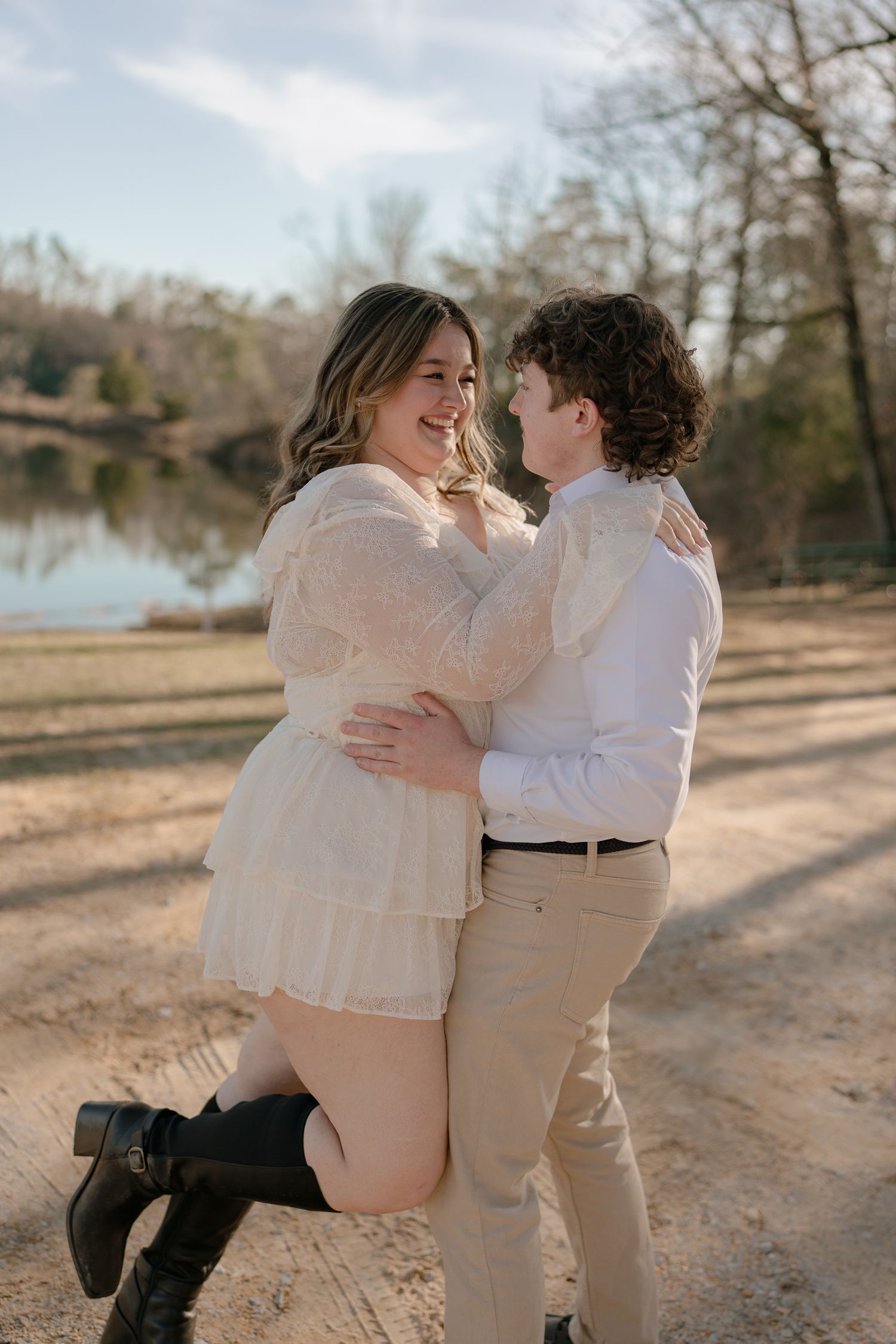 Couple embracing and smiling at each other near a lake, woman in white lace dress and black boots.