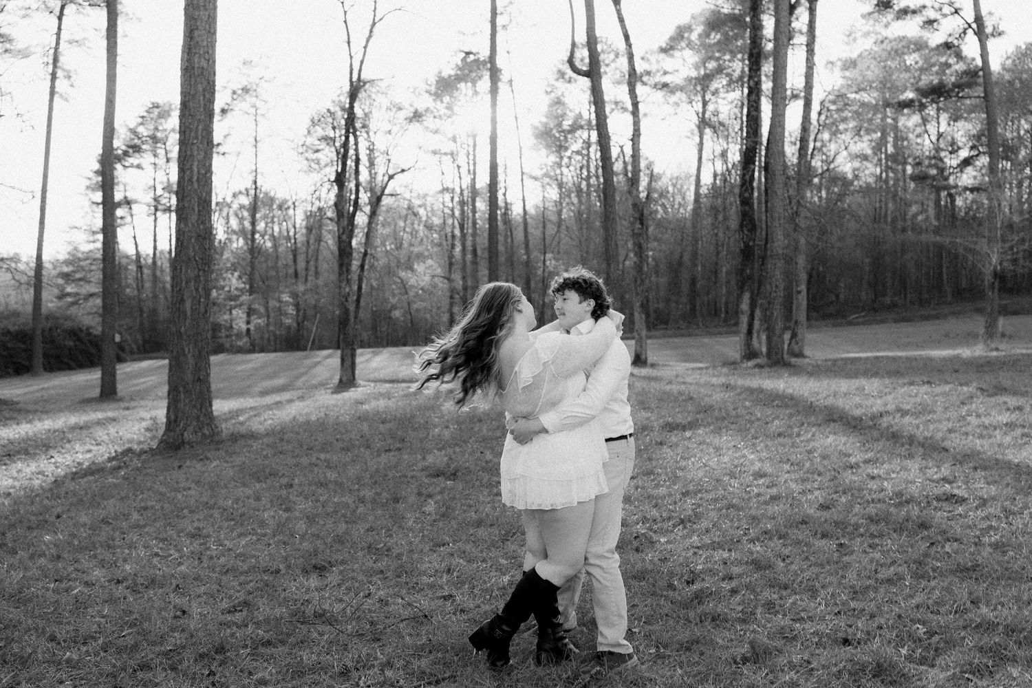 Black and white photo of couple embracing and dancing together in a sunlit forest clearing.