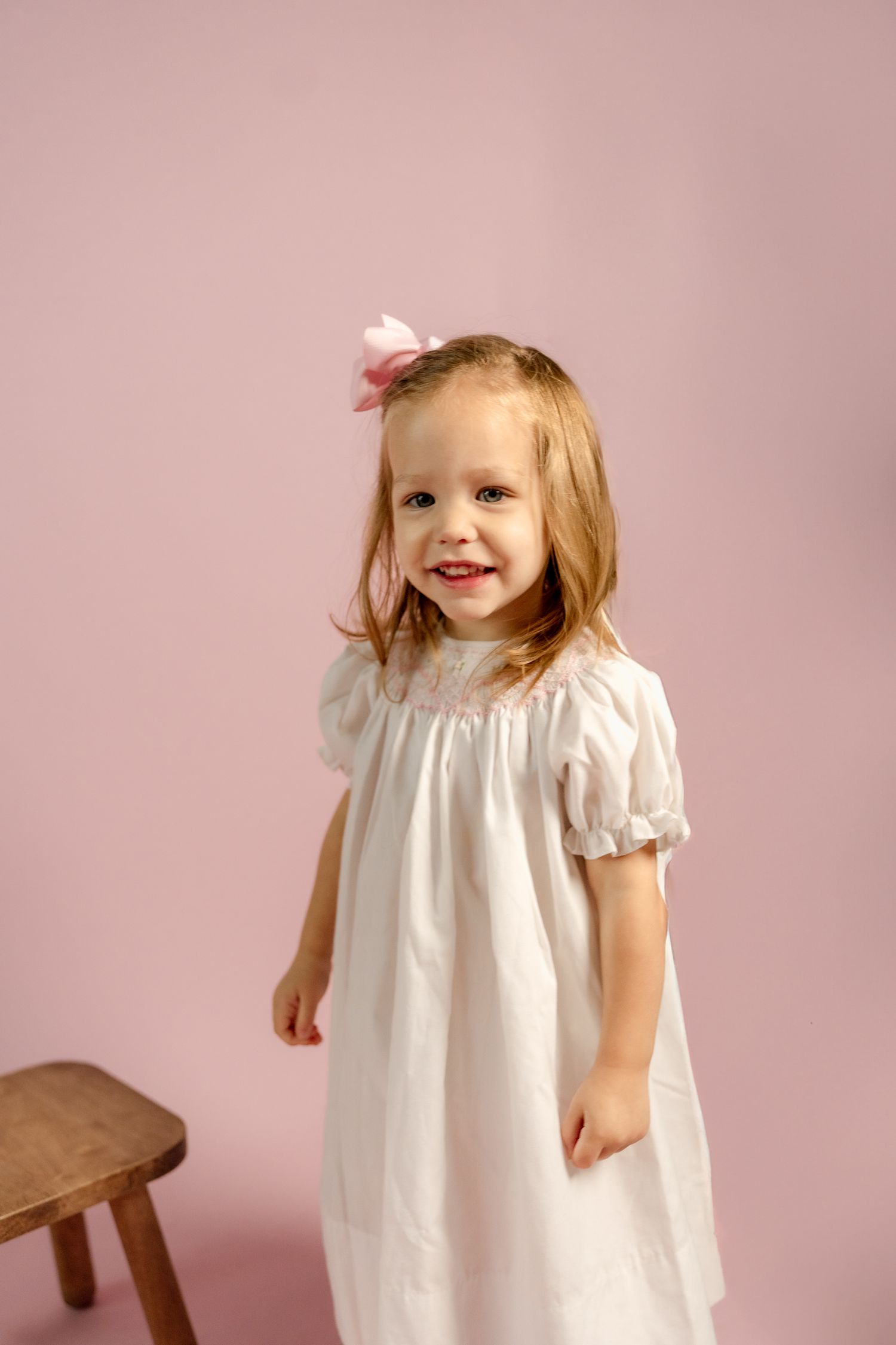 An energetic toddler wearing a white dress poses against a pink studio background.