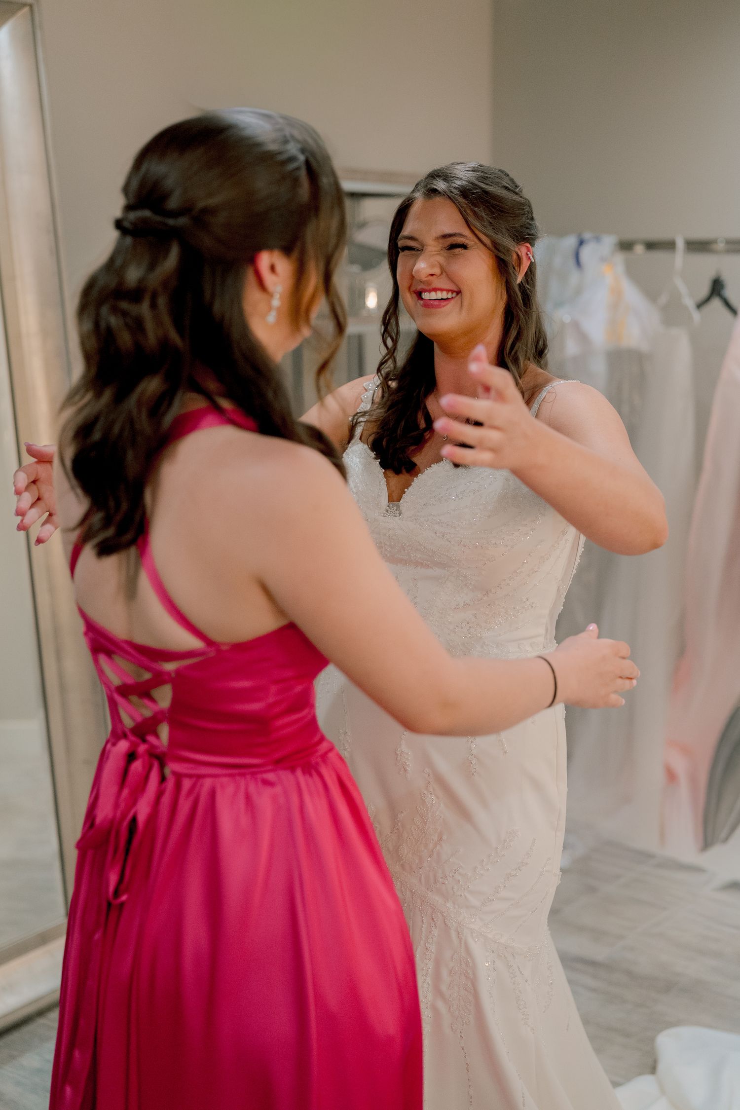 Laughing bride in fitted lace gown sharing joyful moment with bridesmaid in pink satin dress.