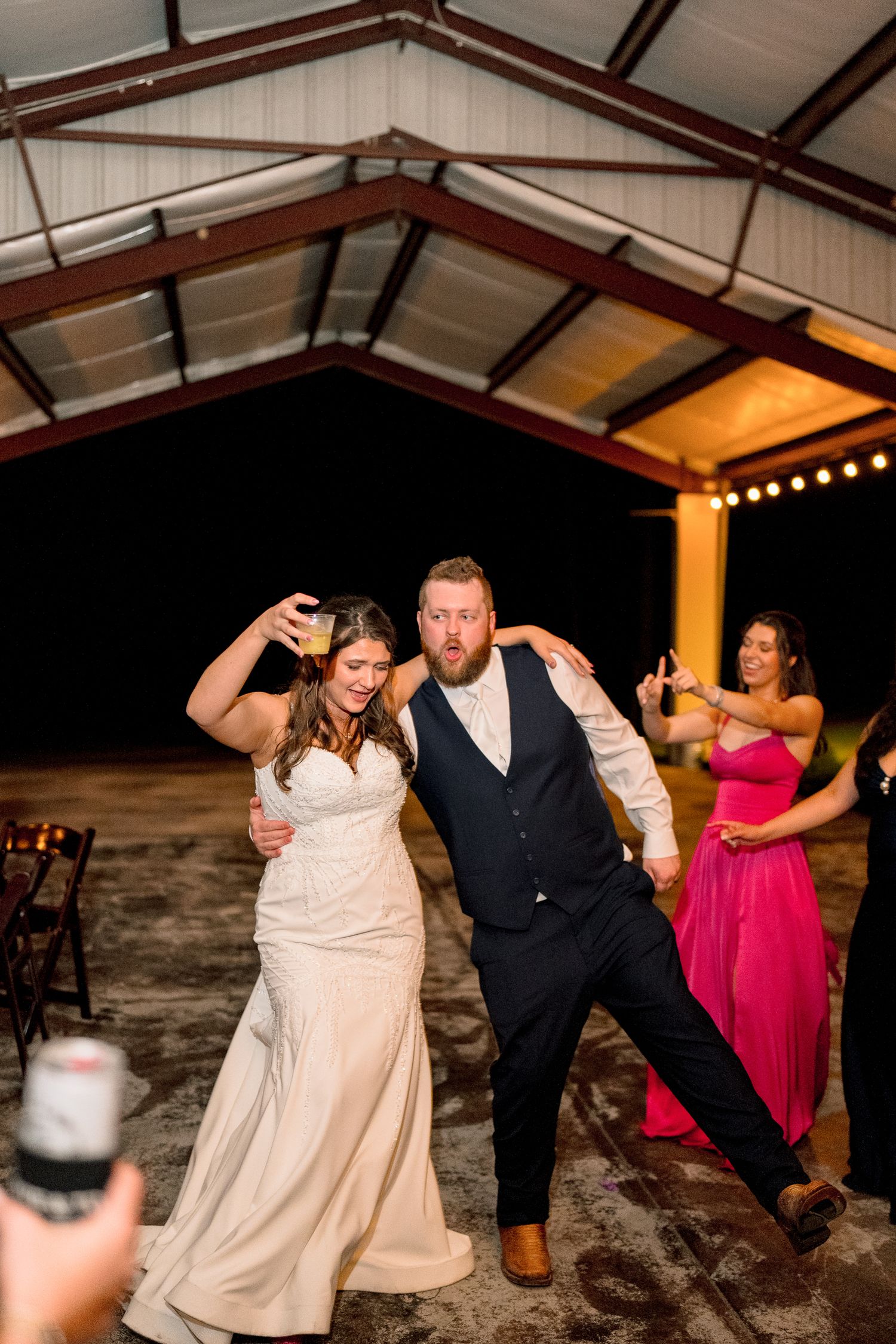 Bride and groom dance joyfully at outdoor evening wedding reception under pavilion with string lights.