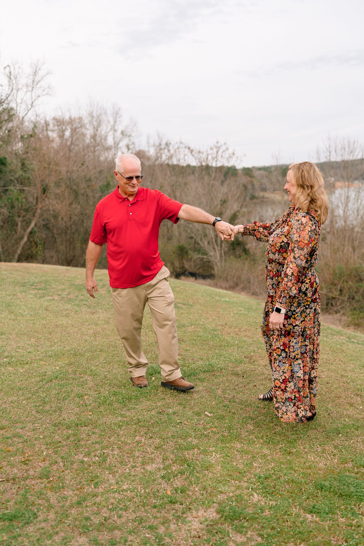Older couple dancing and holding hands on a green lawn with a lake and bare trees in the background.