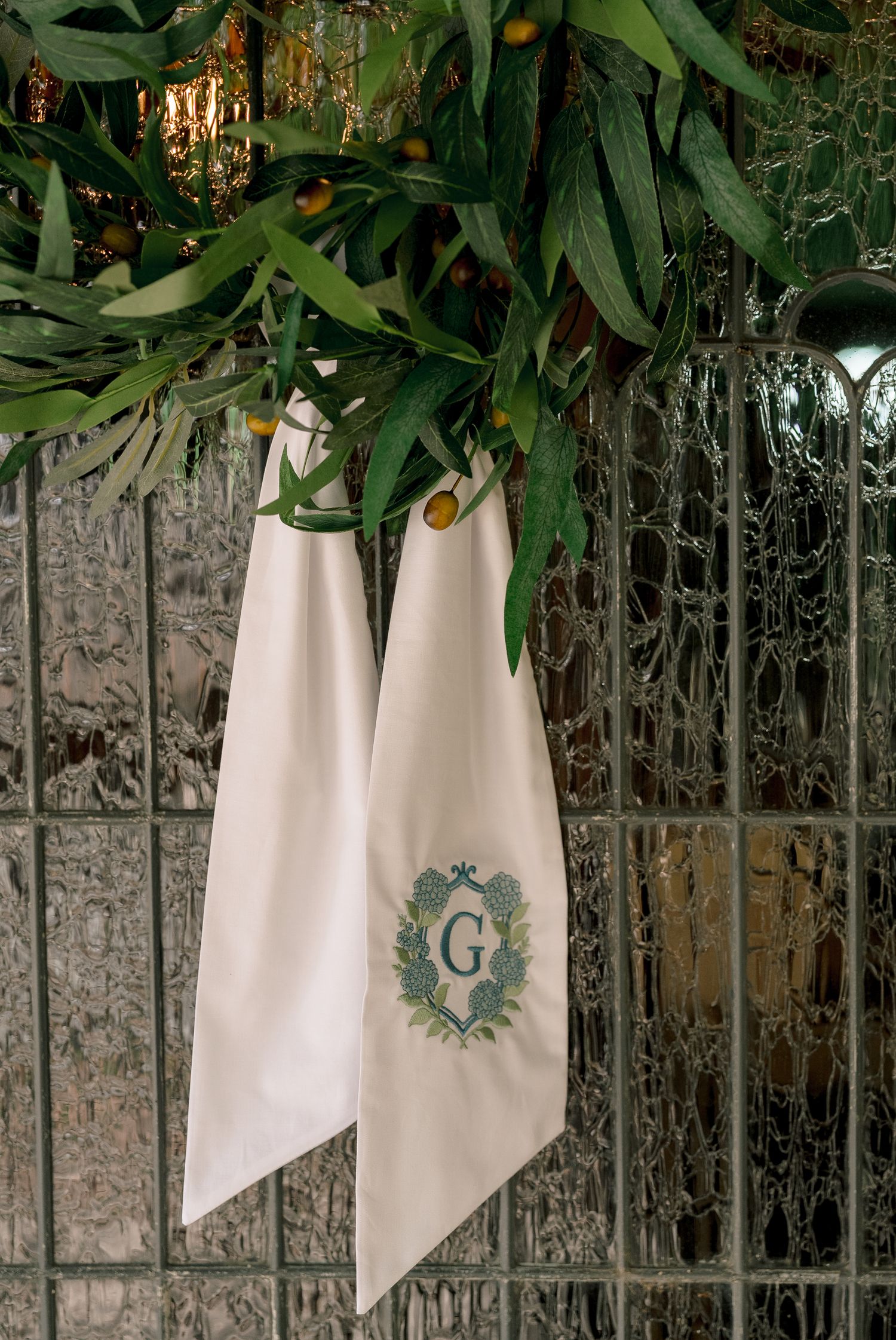White linen napkin with monogram letter G crest hanging from green olive branch garland decoration.