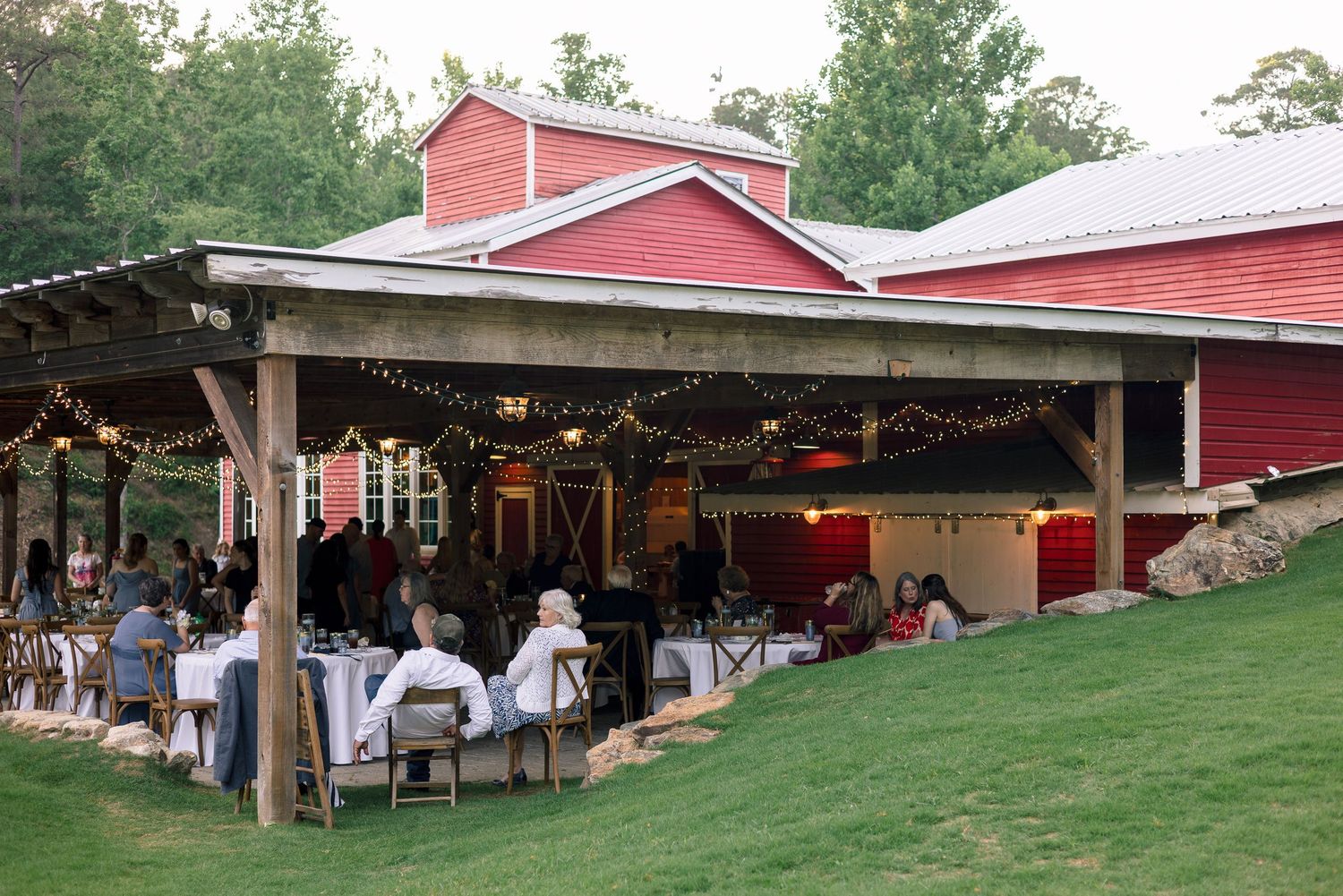 Guests dine at outdoor wedding reception under string lights beside red barn venue at dusk.