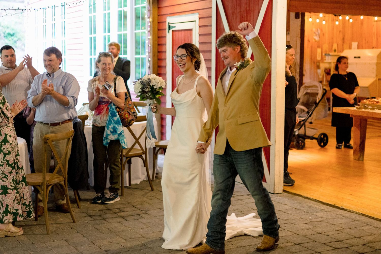 Bride and groom make grand entrance into red barn reception venue as guests cheer and applaud.