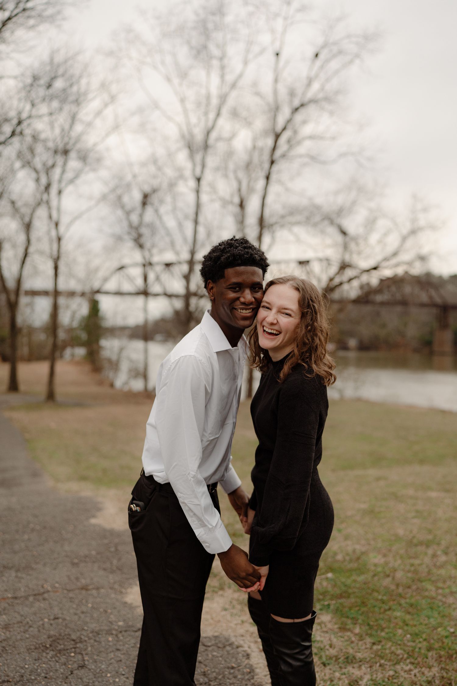 Smiling interracial couple holding hands on a park path surrounded by bare trees.