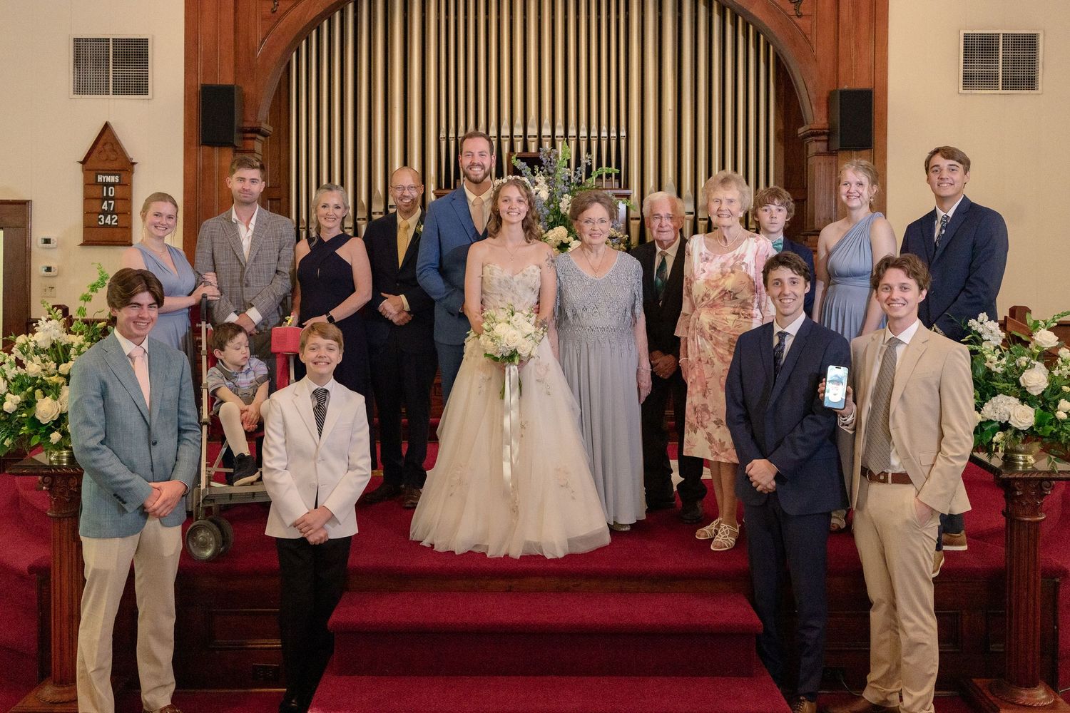 Large wedding party poses together on church altar steps with red carpet and wooden paneling backdrop.