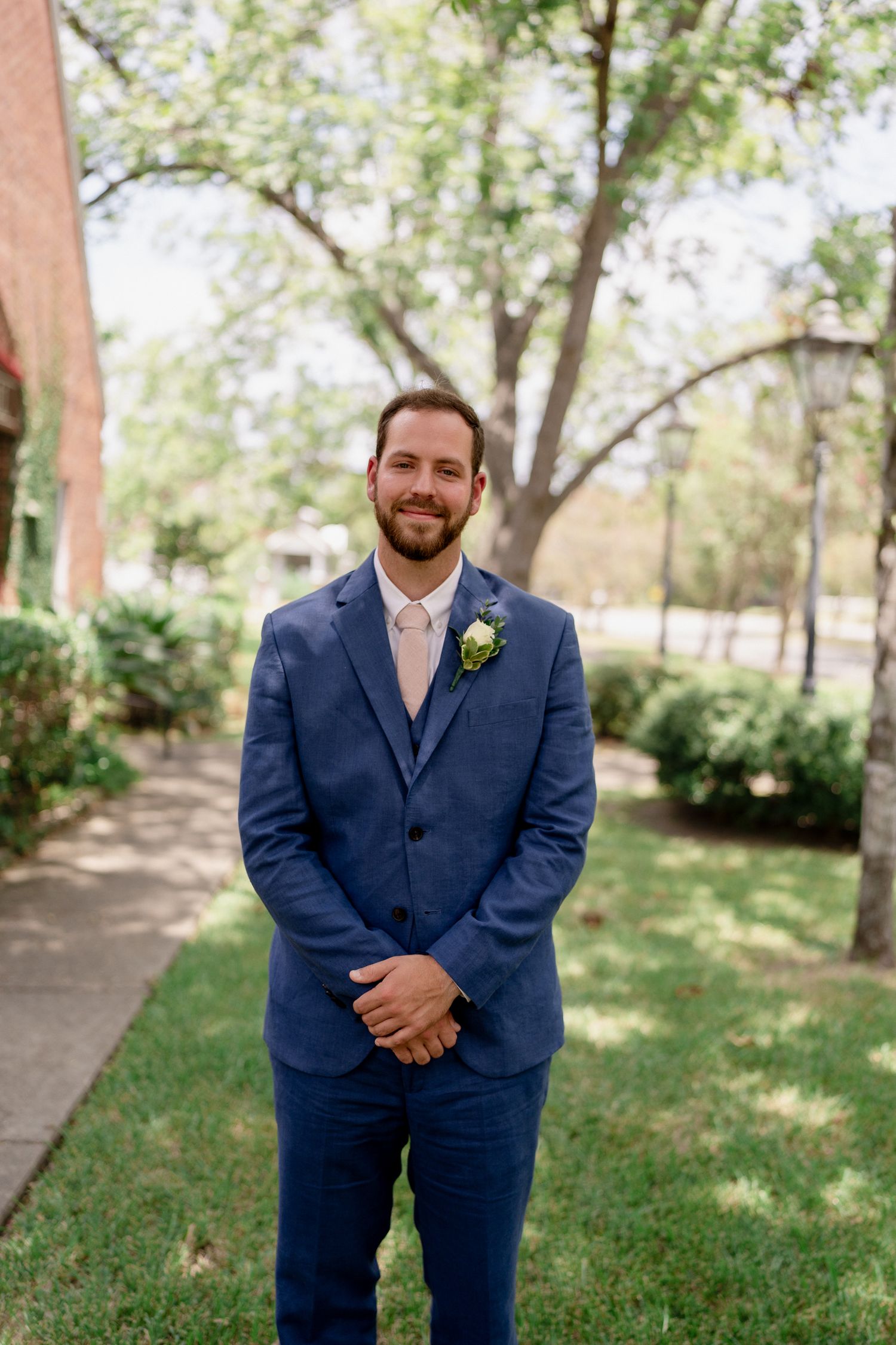 A groom in a navy blue suit poses for portraits outdoors on a sunny day with trees in the background.