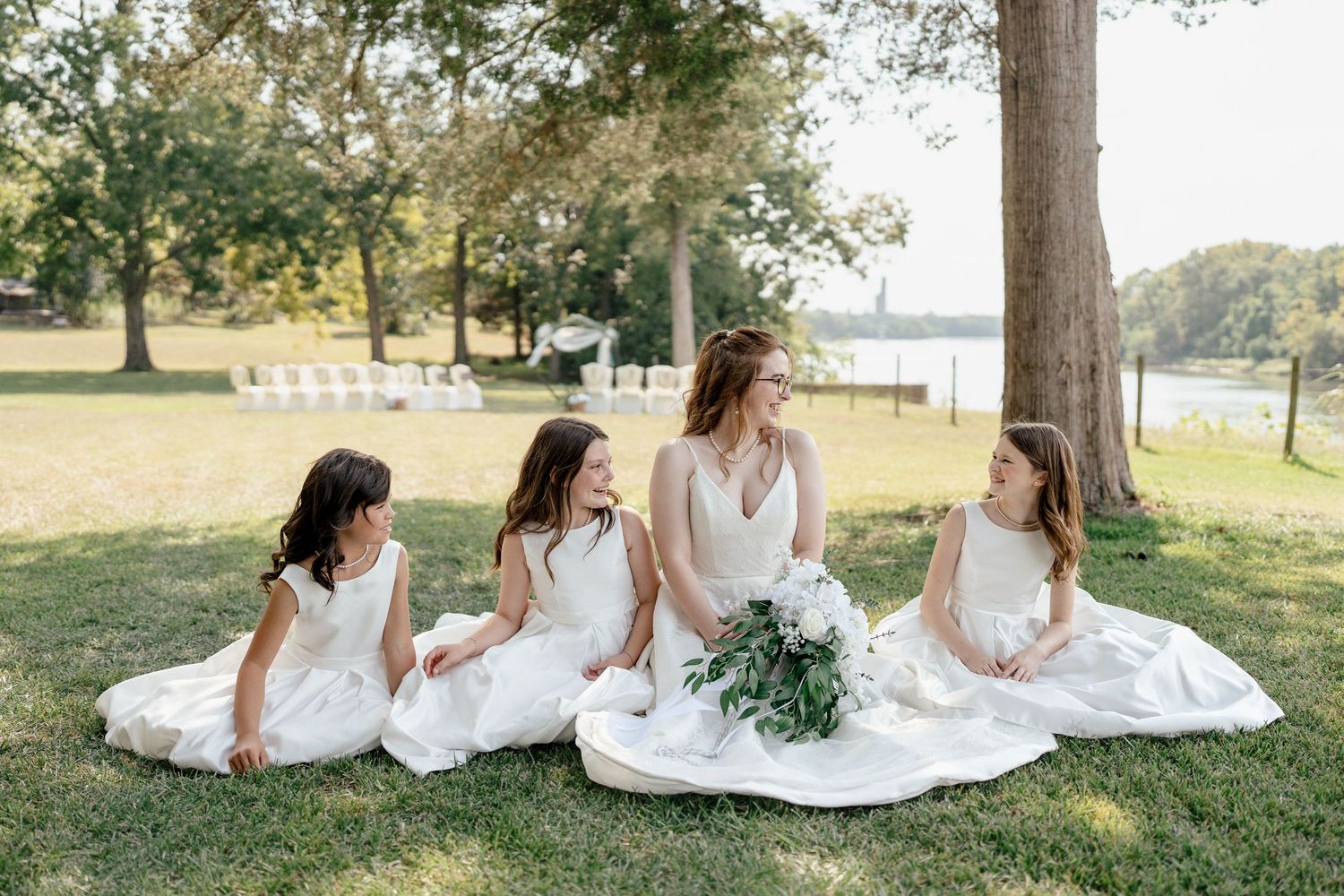 Bridesmaids in white dresses sit together on a grassy hill under a tree.