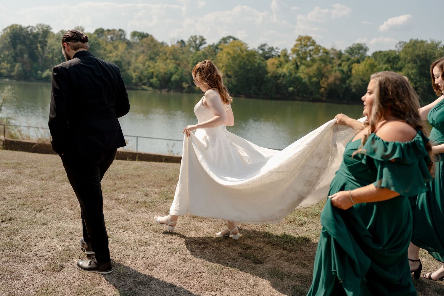 Wedding party in emerald green dresses stands by a lake while a couple poses with flowing white veil in the breeze.