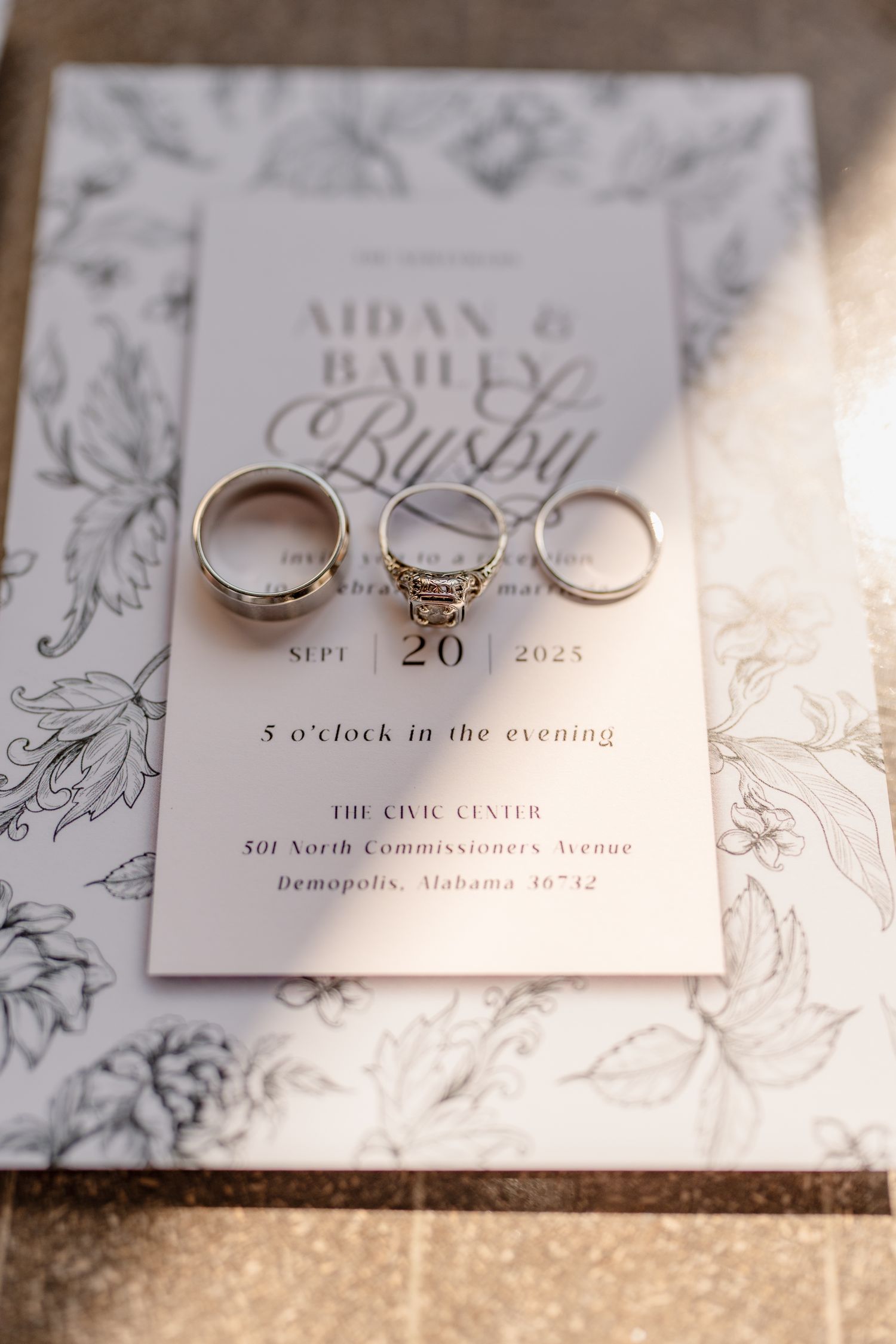Wedding rings displayed on an elegant floral-patterned invitation for Aidan and Bailey's evening ceremony at The Civic Center.
