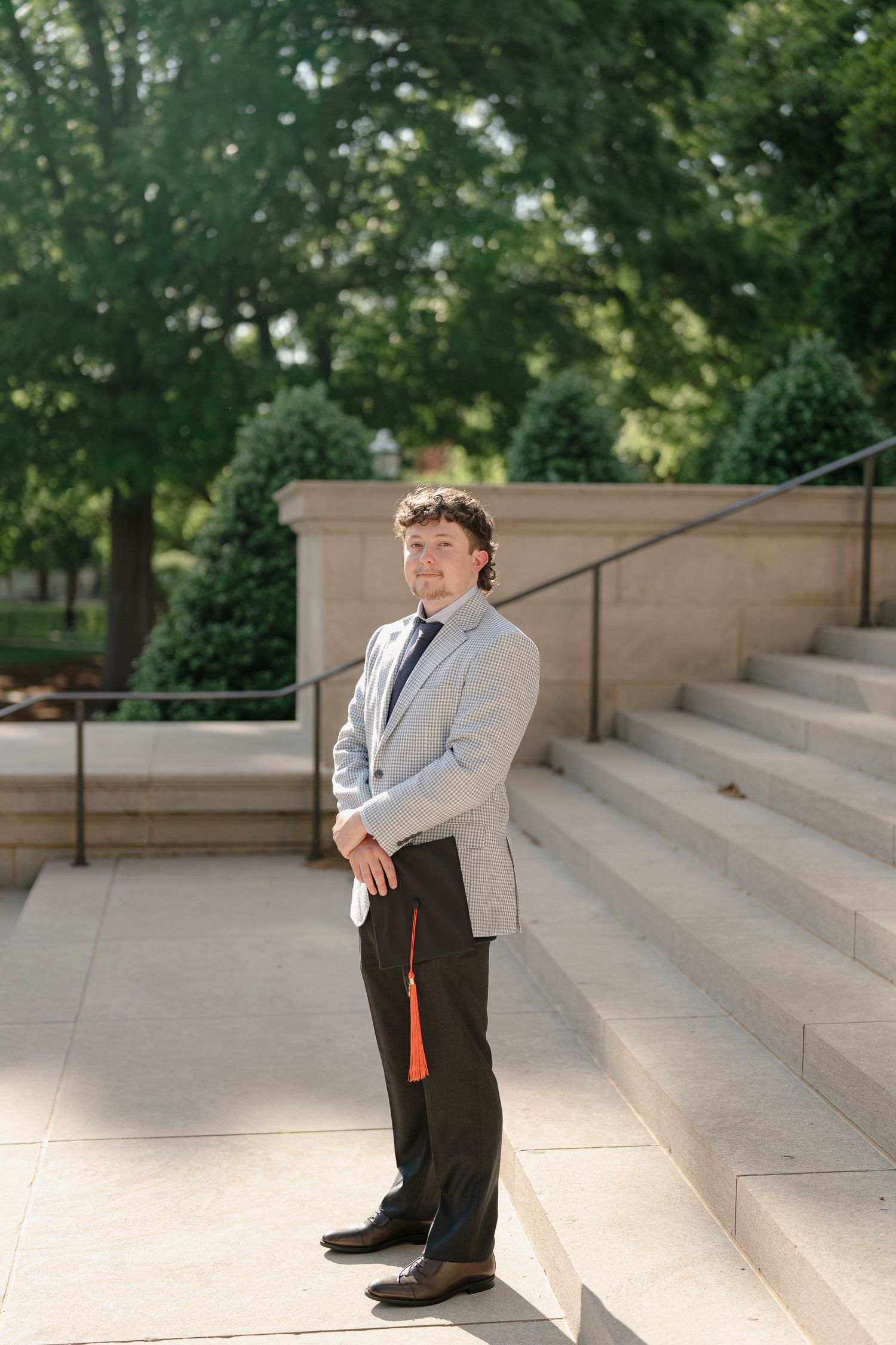 Young man in light gray blazer and dark pants poses confidently on stone steps outdoors near lush green trees.